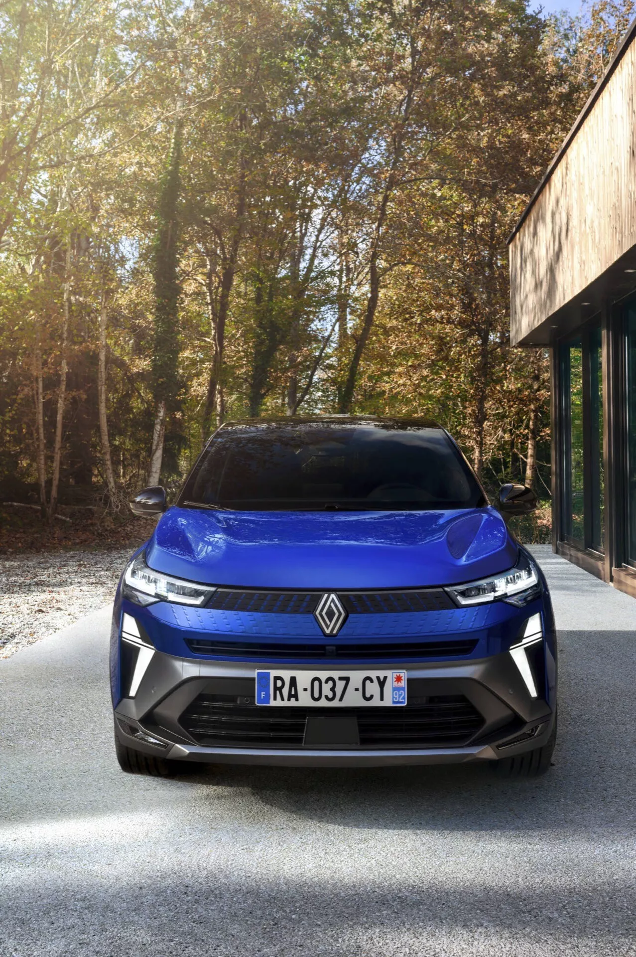 Vista frontal del Renault Captur 2024 con facelift mostrando el diseño de la parrilla, faros LED y emblema en una entrada de asfalto con árboles y un edificio con grandes ventanas de vidrio de fondo bajo condiciones soleadas.