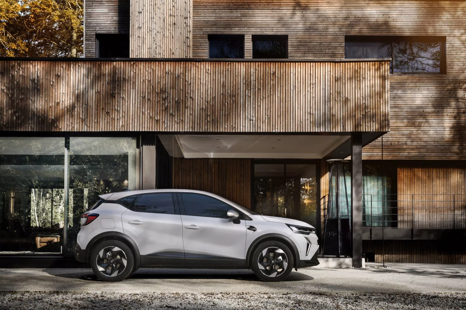 Perfil lateral del Renault Captur II facelift 2024 estacionado frente a una casa moderna con fachada de madera y grandes ventanas de vidrio con elegantes ruedas de aleación negras, diseño elegante de SUV de cuatro puertas y espejos aerodinámicos