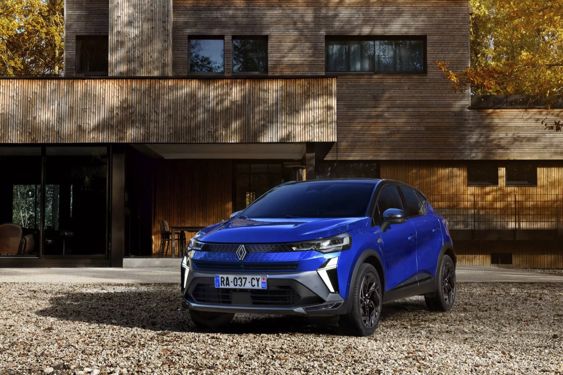 SUV azul Renault Captur (II Facelift) 2024 estacionado en un camino de grava frente a una casa de madera contemporánea, mostrando sus elegantes faros LED, parrilla frontal angular y llantas de aleación intrincadas desde un ángulo frontal bajo
