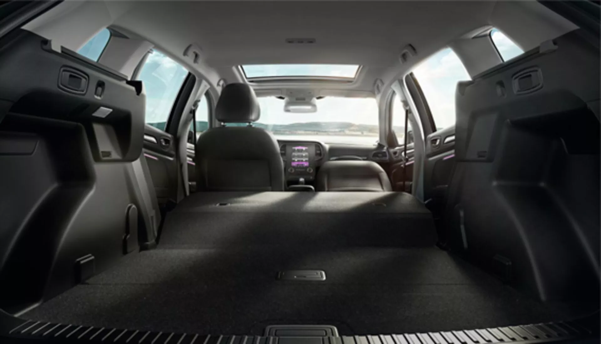 Vista interior del Renault Megane 2016 Grandtour desde la zona de carga hacia el frente, mostrando los asientos traseros abatidos, asientos delanteros tapizados en tela con reposacabezas, bolsillos de almacenamiento en puertas traseras, consola central con portavasos, sistema de infotenimiento iluminado y vista parcial del control del clima y el volante, indicando una cabina espaciosa y equipada con tecnología.