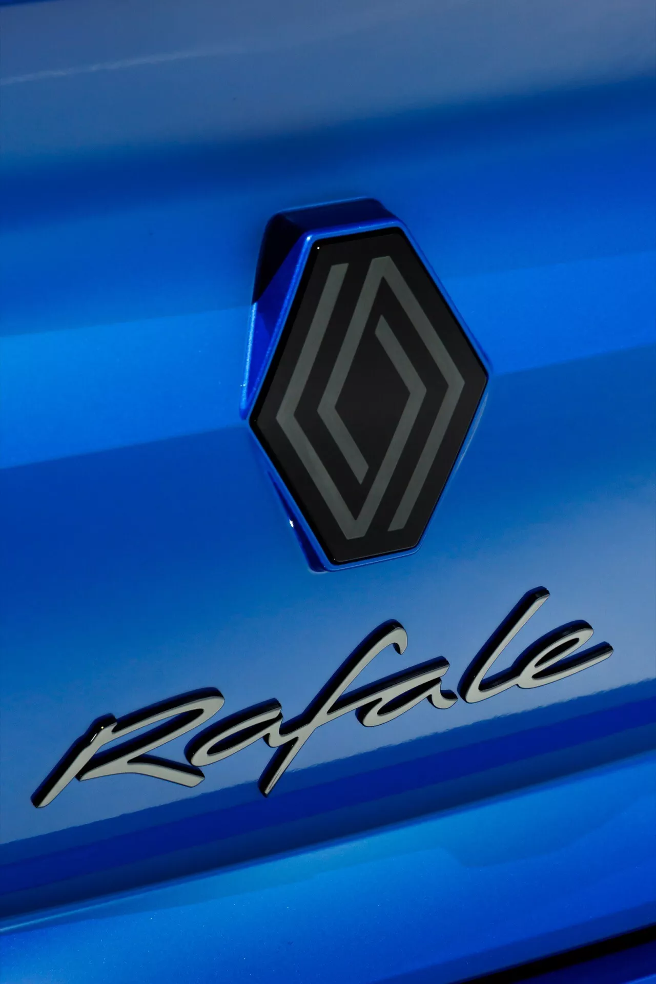 Vista detallada del emblema y nombre del modelo Renault Rafale 2024 (1.ª generación) sobre fondo azul metálico, destacando el diseño hexagonal tridimensional del emblema y la elegante letra 'Rafale'