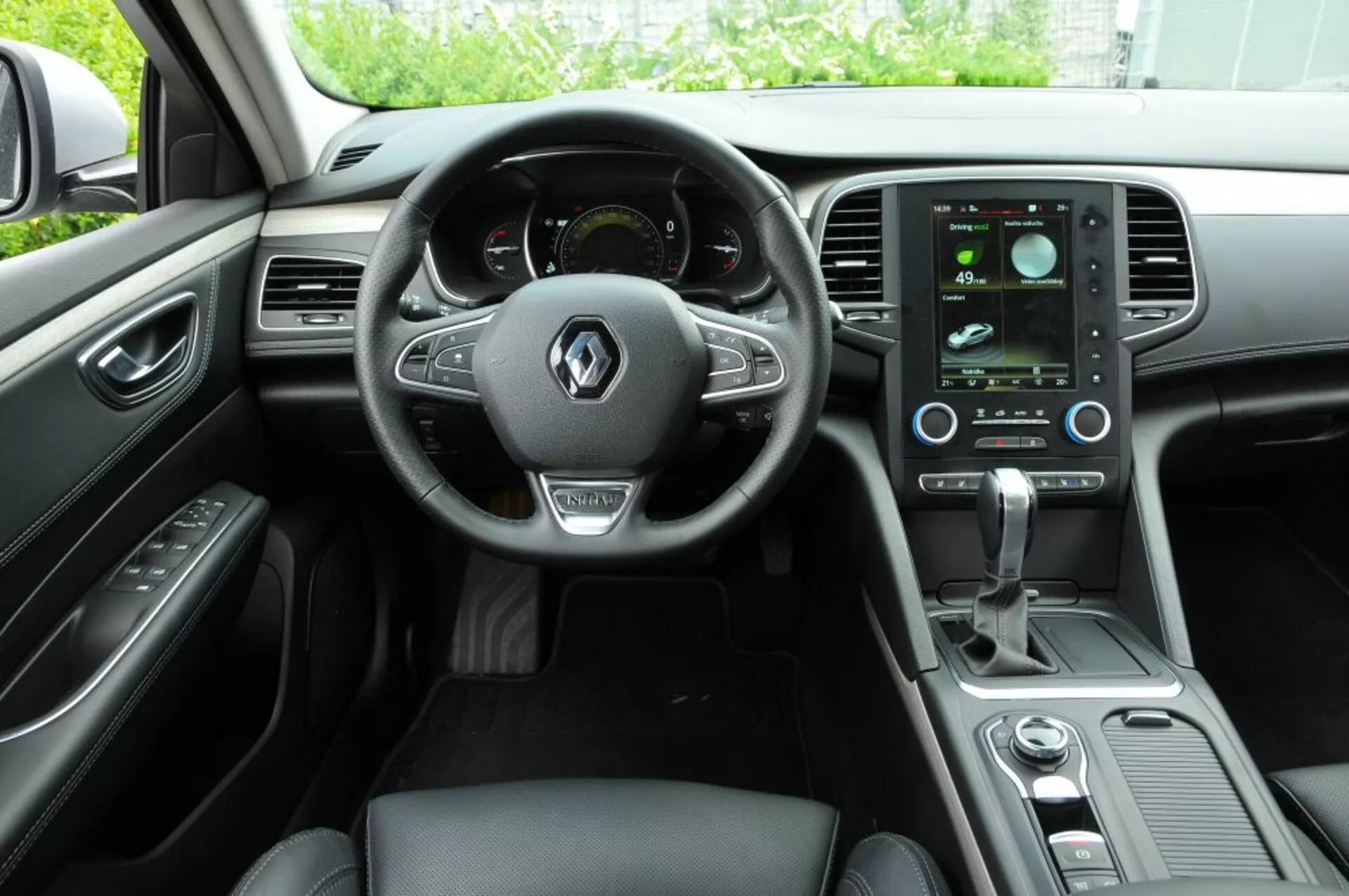 Interior del Renault Talisman 2016 mostrando el cockpit del conductor con volante multifunción de cuero, cuadro de instrumentos digital, sistema de infoentretenimiento táctil vertical y controles de clima elegantes desde la perspectiva del pasajero trasero
