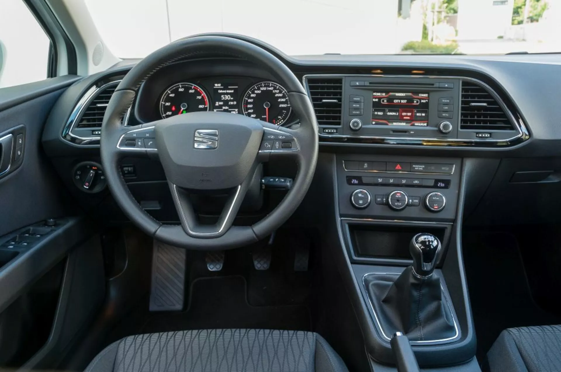 Vista interior de un Seat Leon SC de 2016 desde la perspectiva del asiento del pasajero delantero, mostrando el volante con el logo del fabricante y controles montados, el cuadro de instrumentos digital, el sistema de infoentretenimiento, controles climáticos, el cambio manual, y la puerta del conductor con opciones de energía.