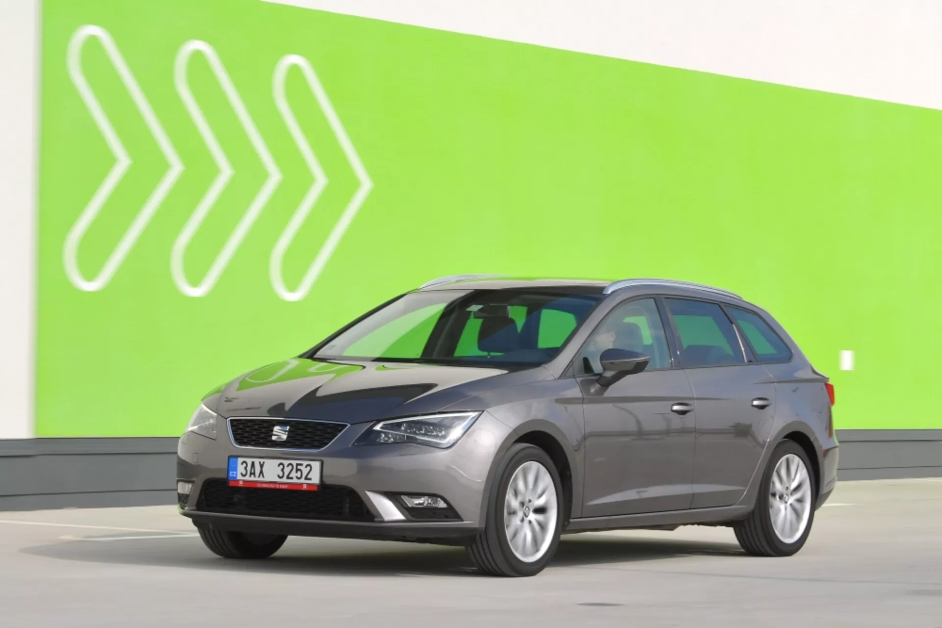 Vagón de estación gris Seat Leon SC 2012 en vista completa de lado contra un fondo verde con cabezas de flecha blancas estilizadas, mostrando espejos laterales con señales de giro, contornos de puertas característicos y ruedas estándar en un suelo de estudio gris claro