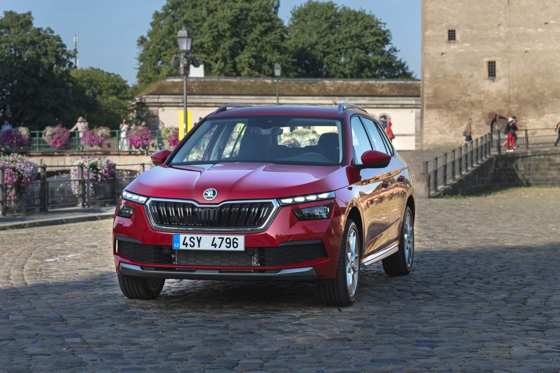 Skoda Kamiq 2019 en rojo, fotografiado desde un ángulo frontal tres cuartos destacando la parrilla frontal, faros LED, capó, parachoques delantero con posibles luces antiniebla o entradas de aire, y llantas de aleación, contra un fondo urbano o histórico de adoquines con follaje.