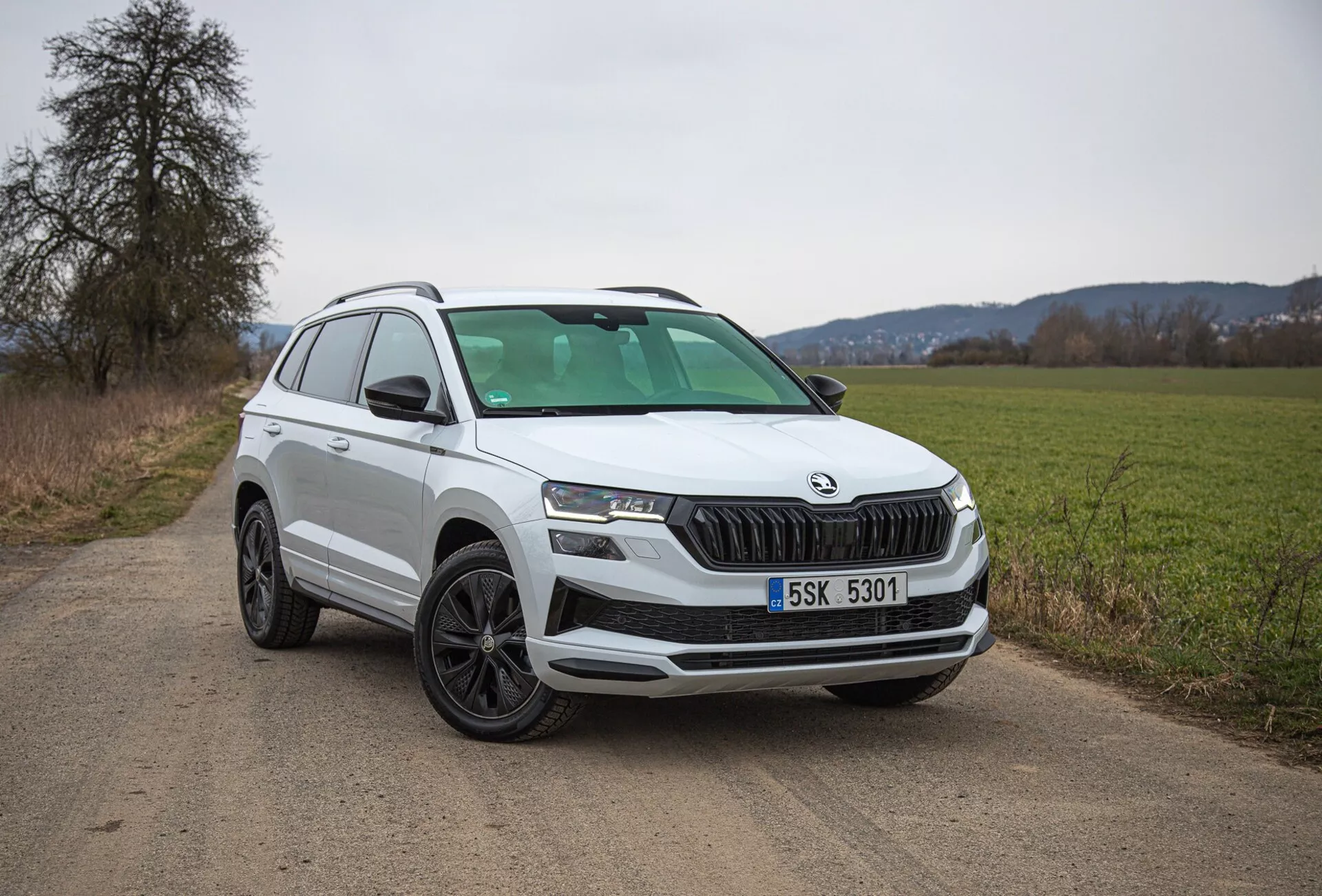 SUV blanco Škoda Karoq de 2021 aparcado en un camino rural de tierra, mostrando vista del cuarto frontal izquierdo incluyendo rejilla, faros y diseño de rueda, con enfoque en características modernas como iluminación LED y sensores de estacionamiento.