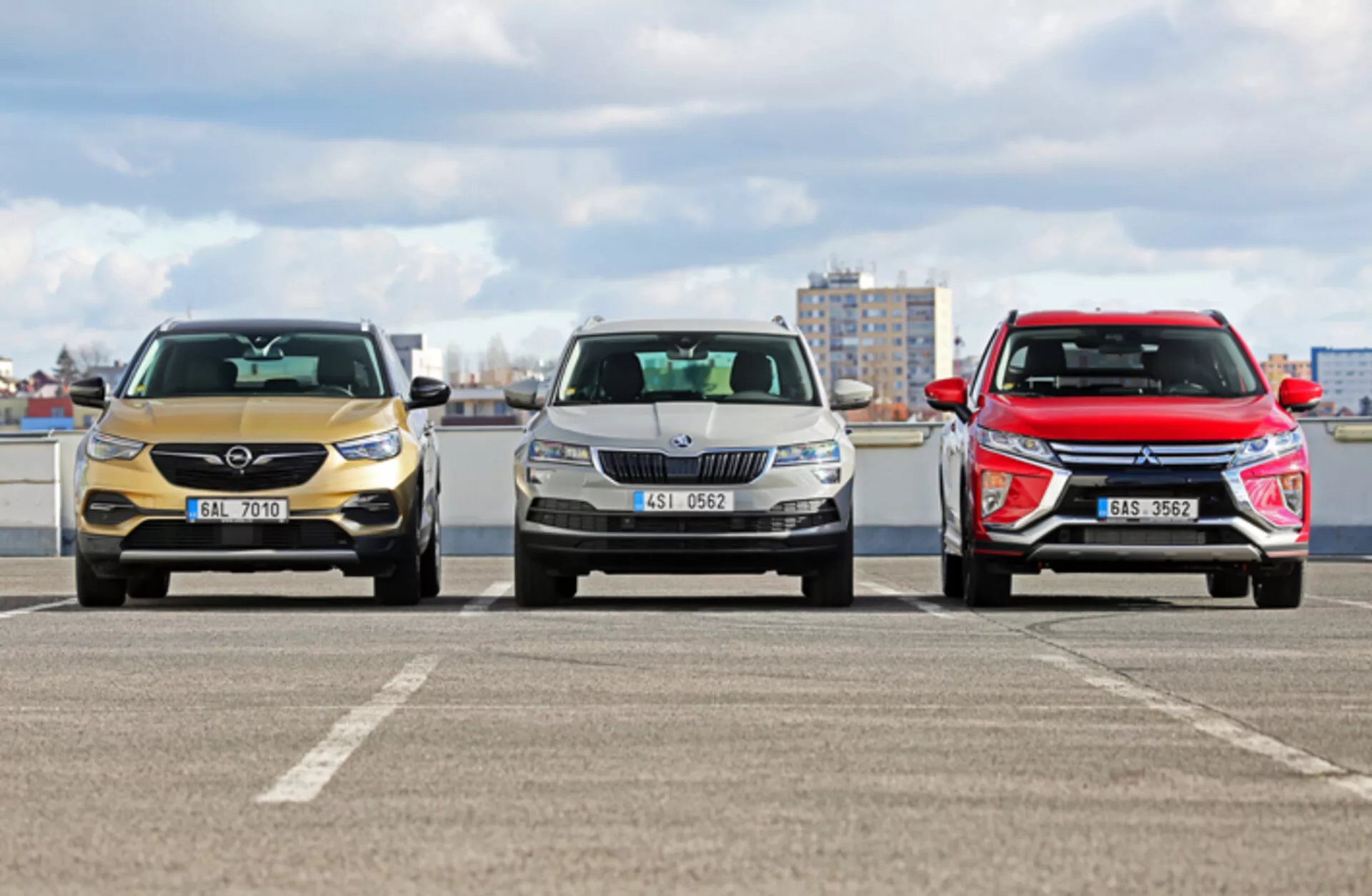 Vista frontal de tres modelos de Skoda Karoq 2017 en un aparcamiento con un paisaje urbano de fondo, mostrando diferentes diseños de parrillas frontales, faros y formas aerodinámicas de capó bajo condiciones de luz diurna nublada