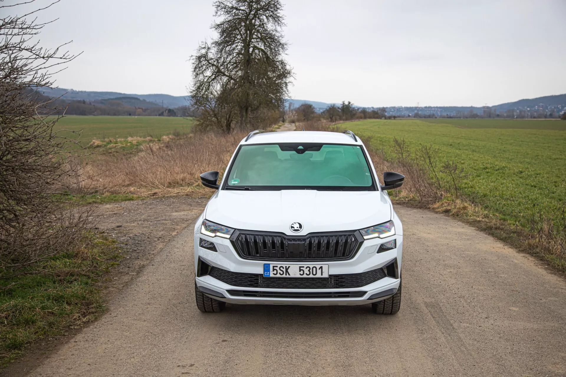 SUV Škoda Karoq 2021 mostrando una vista frontal directa sobre un camino rural estrecho y sin pavimentar, destacando la distintiva parrilla de Škoda, faros LED afilados, luces de niebla dentro del borde negro para capacidad todo terreno potencial, líneas dinámicas del capó y características tecnológicas avanzadas en el frontal del vehículo.