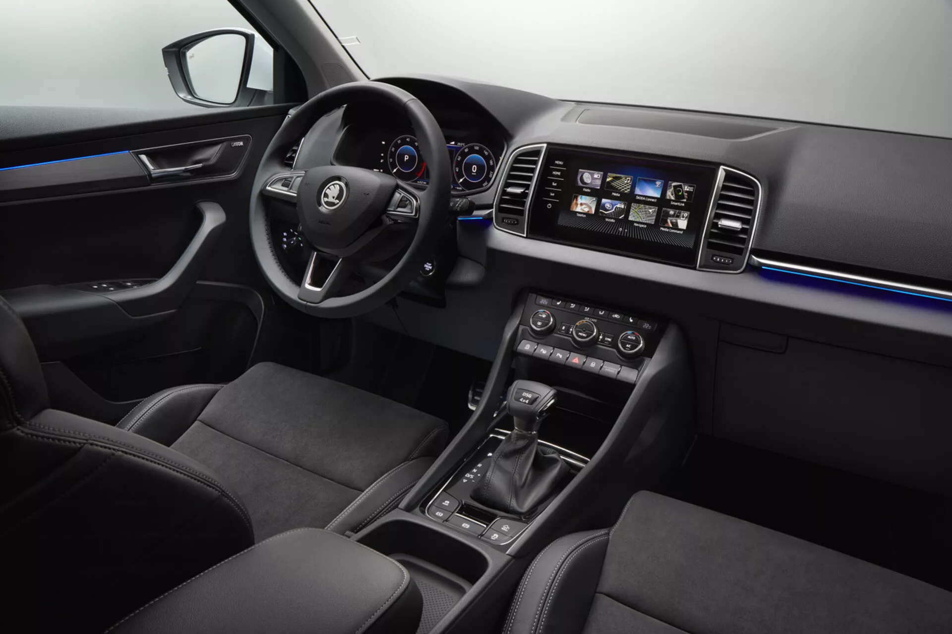 Vista interior del Skoda Karoq 2017 mostrando el lado del conductor con enfoque en el volante con logo, tablero digital, consola central iluminada con sistema de infotenimiento táctil, control climático automático, palanca de cambios con selectores de modo de conducción, iluminación ambiental azul y asientos de tela oscura de calidad premium.