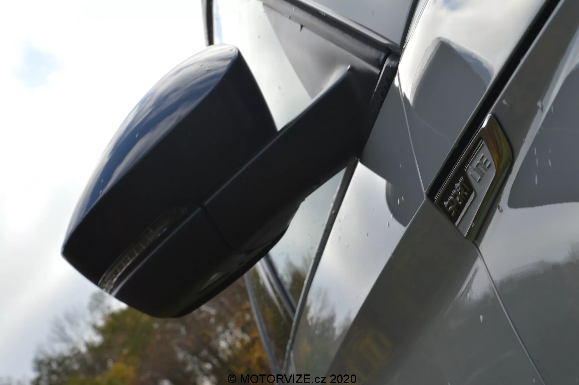 Vista lateral en primer plano de un Skoda Kodiaq 2017 centrada en el espejo exterior y la zona del guardabarros. La imagen está tomada desde un ángulo bajo, enfatizando el perfil lateral del coche, con una carcasa de espejo lateral moderna y aerodinámica a juego con el color del vehículo, una insignia prominente en el guardabarros que indica un modelo o nivel de acabado específico, paneles de carrocería metálicos y brillantes, y reflejos que sugieren un entorno al aire libre con un coche de color oscuro.