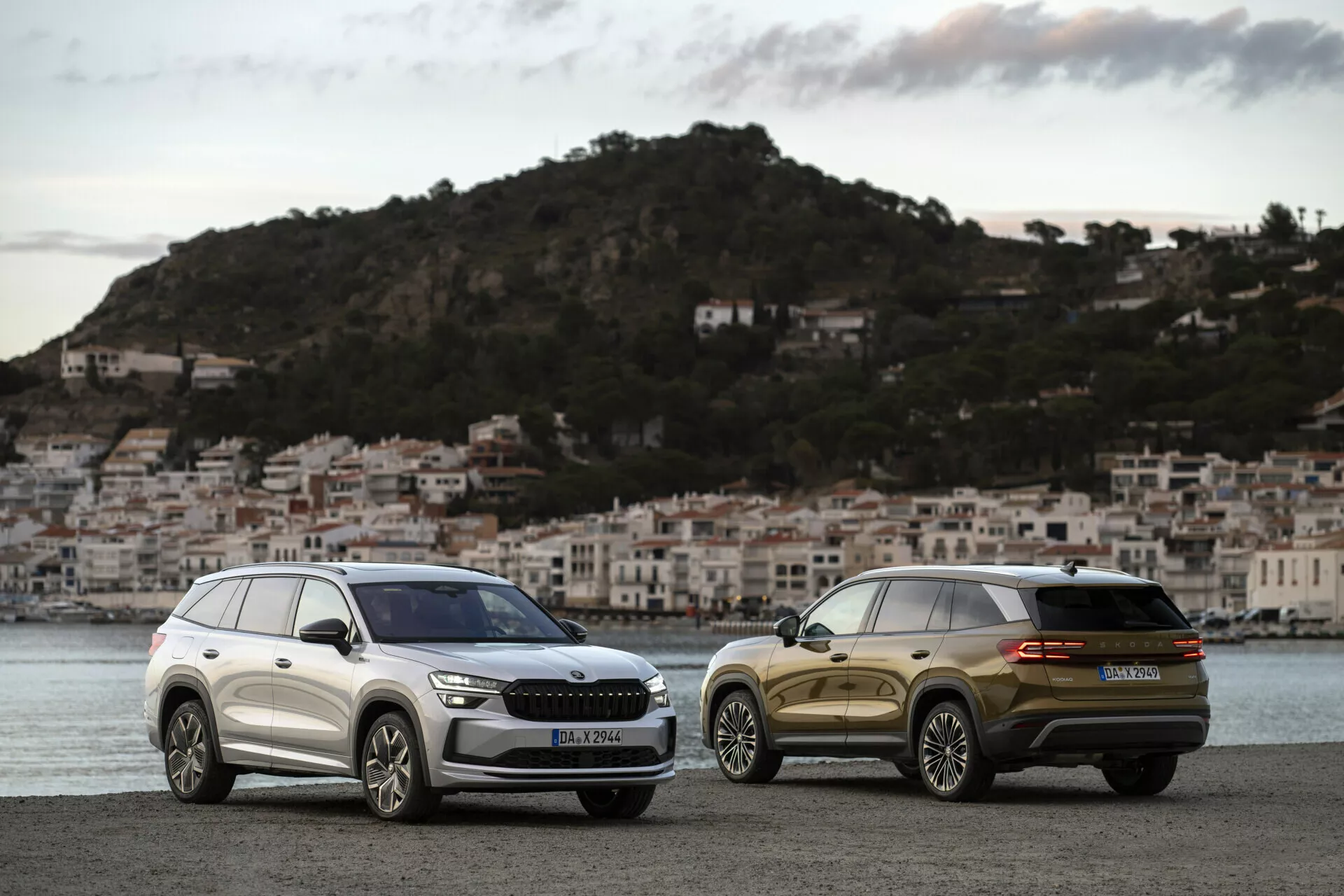 SUVs Skoda Kodiaq 2024 estacionados uno al lado del otro al amanecer en un paseo marítimo con un pueblo costero de fondo, mostrando la parrilla frontal, faros y líneas de diseño elegantes de uno y la vista trasera de tres cuartos del otro.