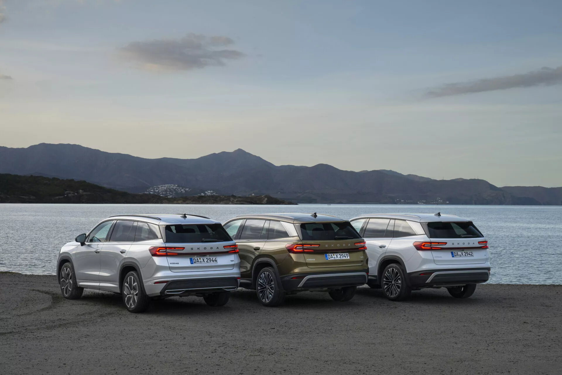 Alineación de Skoda Kodiaq 2024 con tres SUV estacionados en la orilla del agua al crepúsculo, mostrando perfiles traseros y estética de diseño contra un fondo de paisaje suavemente iluminado