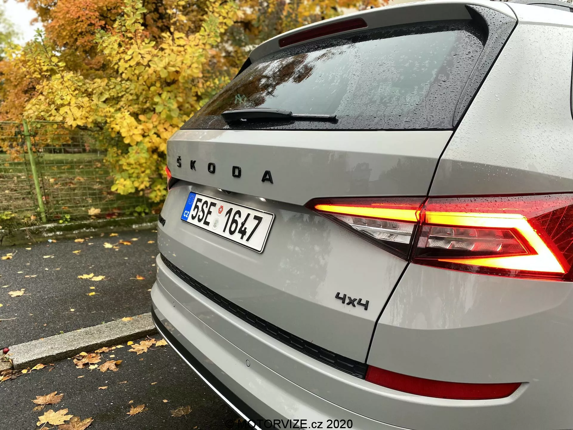 Vista trasera de un vehículo plateado Škoda con luces traseras LED en forma de C iluminadas, emblema y marca Škoda, con insignia 4x4, capturado desde el lado trasero derecho sobre un pavimento mojado entre hojas de otoño y árboles.