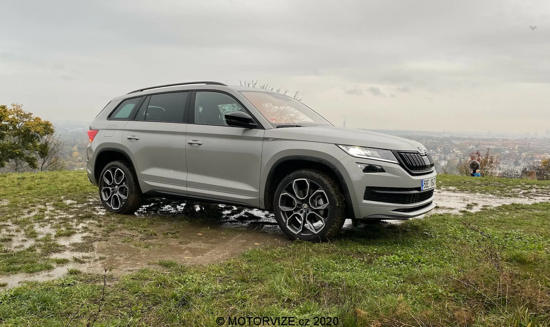SUV Skoda Kodiaq plateado de 2017 aparcado en suelo de césped ligeramente embarrado, captado desde un ángulo delantero lateral. Vista clara de la parrilla frontal, faros, perfil lateral izquierdo, diseño de faros elegantes con tecnología LED, diseño distintivo de la parrilla, posibles sensores de sistemas de asistencia bajo la parrilla, espejos laterales con indicadores y posible detección de puntos ciegos, ruedas de aleación multirradio grandes, neumáticos adecuados para condiciones en carretera y moderadamente fuera de ella, sugiriendo capacidades de tracción total. Posicionado en una pendiente con paisaje urbano nebuloso en el fondo bajo un cielo nublado, luz suave que indica temprano por la mañana o tarde por la tarde. Personas en el fondo disfrutando de la vista, destacando el atractivo recreativo y de estilo de vida.