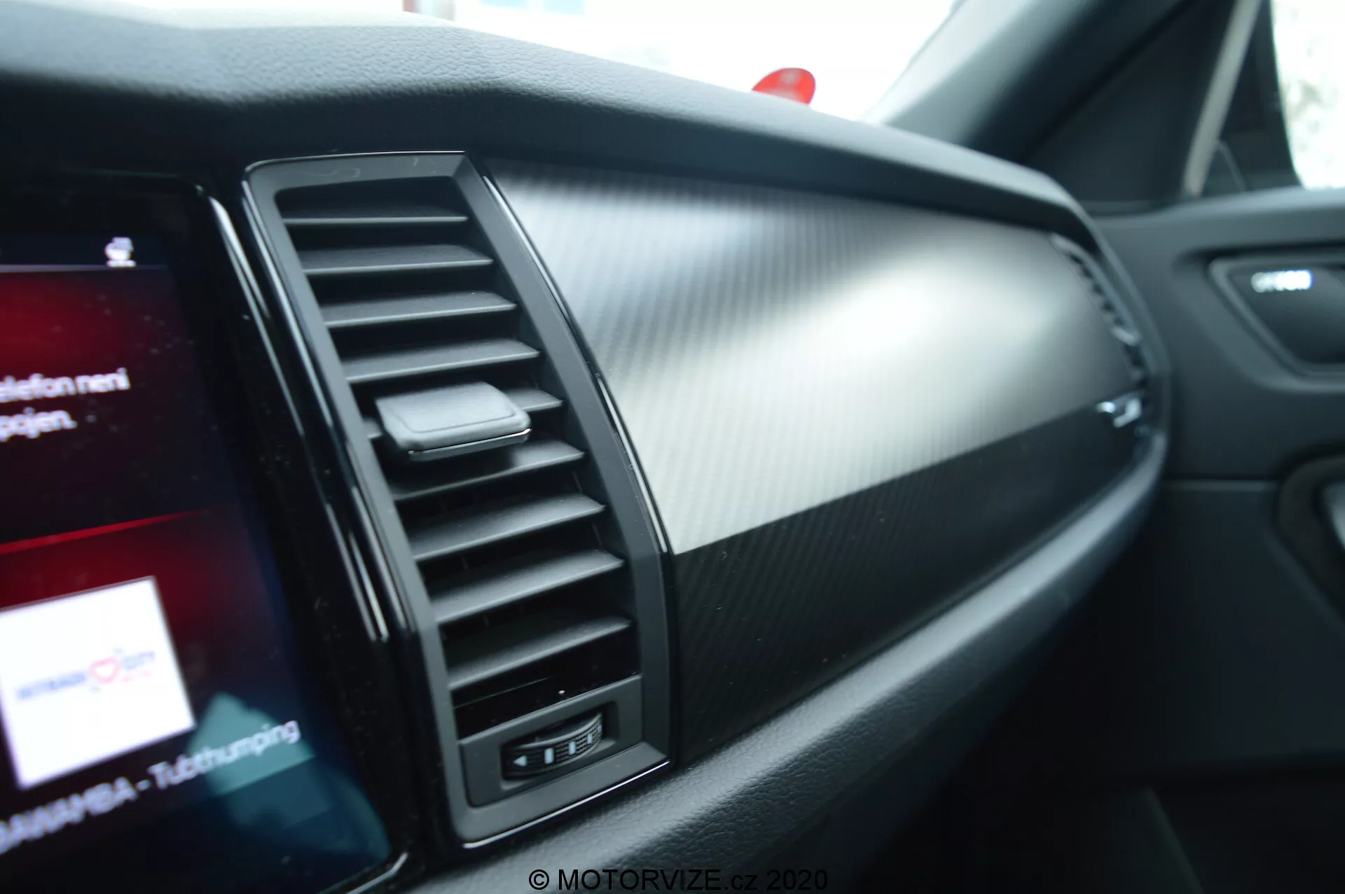 Vista de cerca del interior del Skoda Kodiaq 2017, destacando el lado del pasajero del tablero. Características incluyen una ventilación de aire con lamas ajustables verticalmente, una sección de trim decorativo y una pantalla de infoentretenimiento parcialmente visible con gráficos coloridos. Los materiales del tablero combinan metal cepillado y plástico texturizado que se asemeja al carbono, enfatizando un diseño moderno y deportivo.