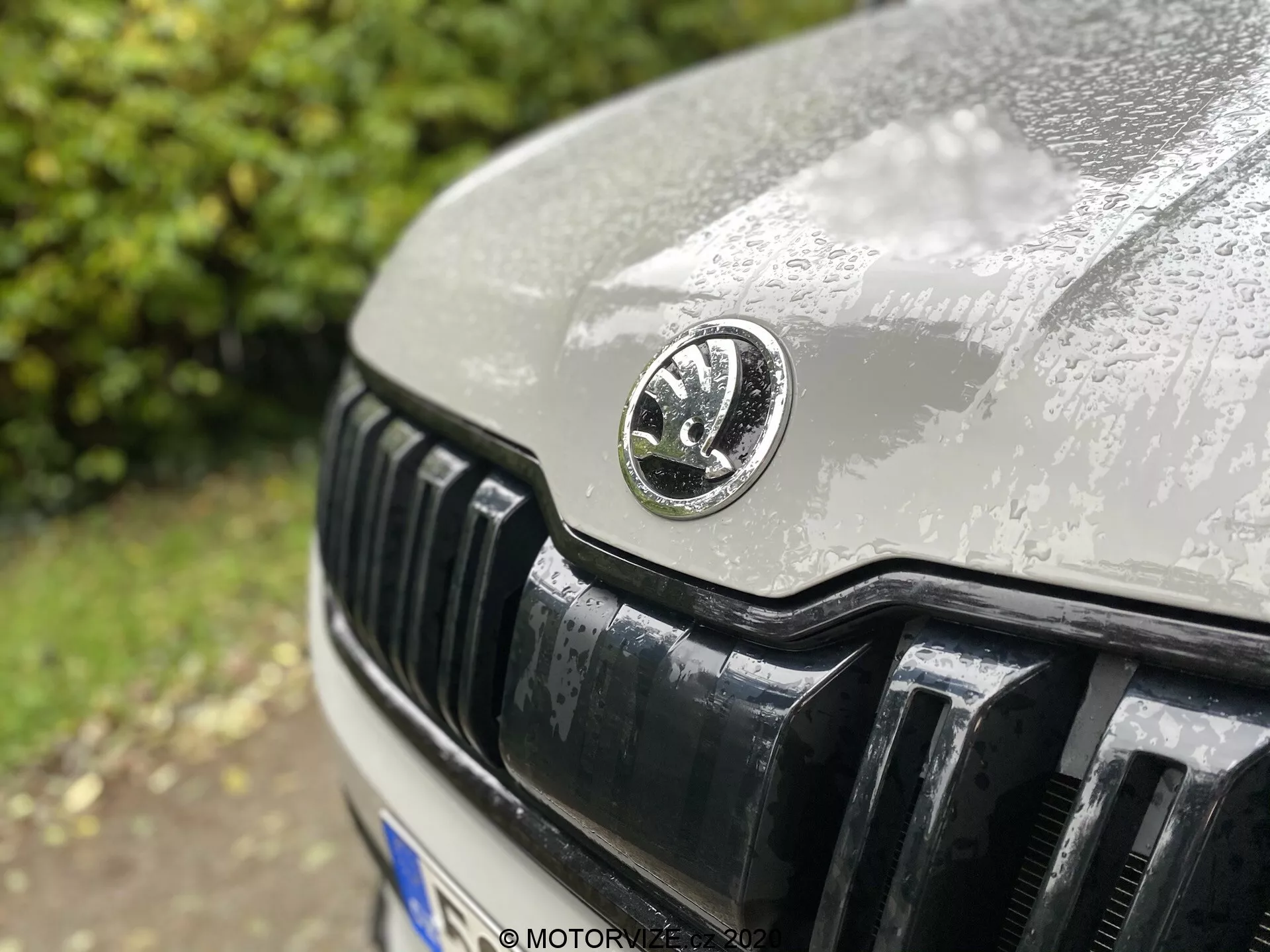 Primer plano delantera del Skoda Kodiaq 2017 enfocado en el emblema de Skoda, mostrando la rejilla negra brillante con láminas verticales, el capó cubierto de gotas de lluvia, y parte del faro derecho, con fondo desenfocado.