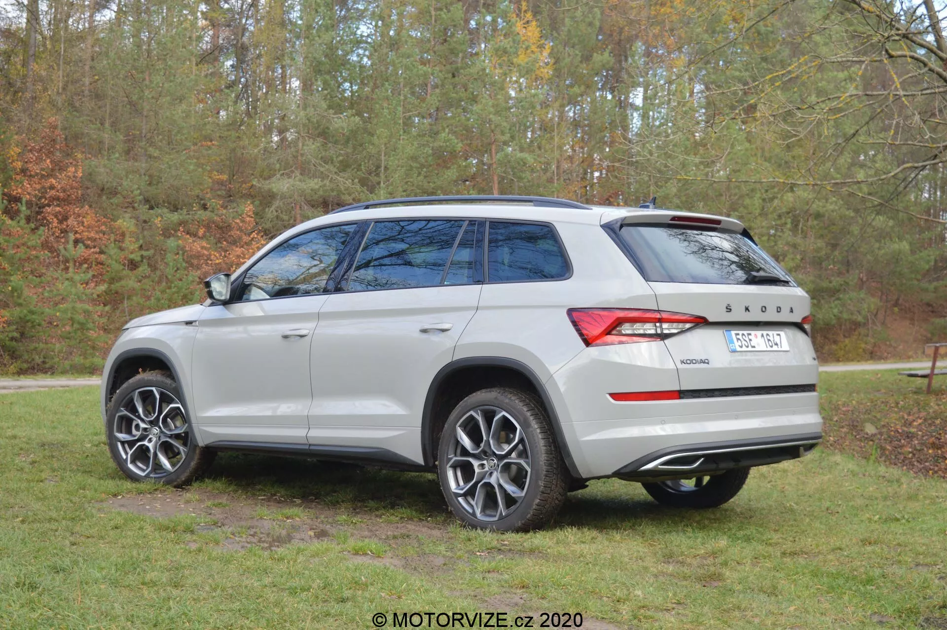 Imagen de un Škoda Kodiaq 2017 capturada desde un ángulo de tres cuartos trasero, que muestra el color plateado/gris claro del vehículo, las llantas de aleación de múltiples radios, las ventanas traseras tintadas, el techo solar panorámico y el diseño distintivo de las luces traseras. El automóvil está estacionado al aire libre con un fondo de árboles y hierba, lo que indica un entorno natural. En el portón trasero son visibles los distintivos de Škoda y el emblema de la tracción '4x4'.