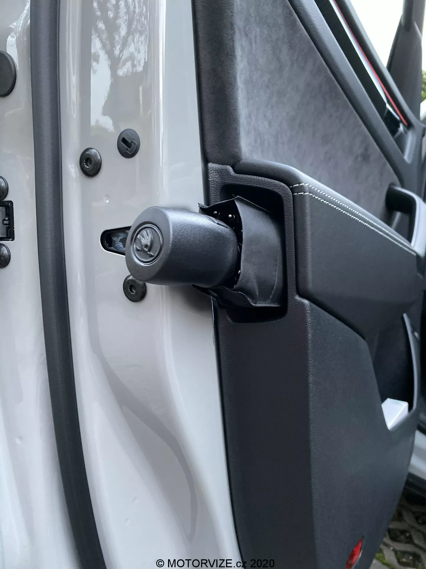 Vista de primer plano del robusto mecanismo de bisagra de la puerta y el área circundante en un Škoda Kodiaq 2017, con un componente de resorte visible para uso pesado. El vehículo blanco contrasta con los componentes de bisagra negros, incluido un mecanismo de cerradura tradicional con orificio para llave encima de la bisagra, asegurado con pernos de cabeza hexagonal para durabilidad. El plástico texturizado del panel interior de la puerta y los puntos de montaje seguros mejoran el enfoque del diseño utilitario. Los elementos de fondo desenfocados resaltan el mecanismo de bisagra y cierre.