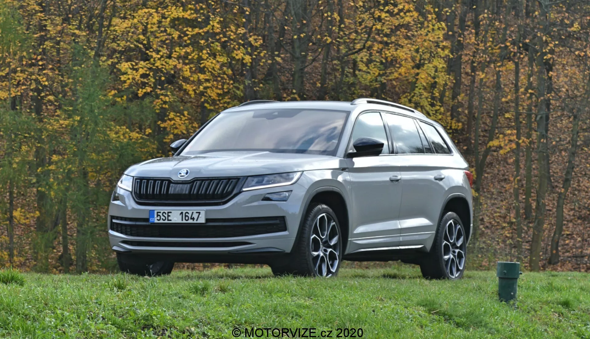 La imagen muestra un SUV Skoda Kodiaq 2017 estacionado en un área con césped y árboles de otoño en el fondo. Fotografiado desde una vista frontal de tres cuartos, el vehículo presenta una rejilla frontal de lamas horizontales con el logotipo de Skoda en el centro. Los faros LED elegantes se extienden hacia los lados, acompañados por luces antiniebla en la parte inferior del parachoques. El capó tiene líneas sutiles y un borde plateado adorna la parte inferior del parachoques frontal. Ruedas de aleación con diseño multirradio, espejos laterales con señales de giro integradas, rieles en el techo y líneas esculpidas destacan el perfil. Los sensores o cámaras en la parte inferior del parachoques sugieren avanzados sistemas de asistencia al conductor (ADAS). El entorno al aire libre incluye una carretera asfaltada y un contenedor de residuos verde en el borde derecho de la imagen.