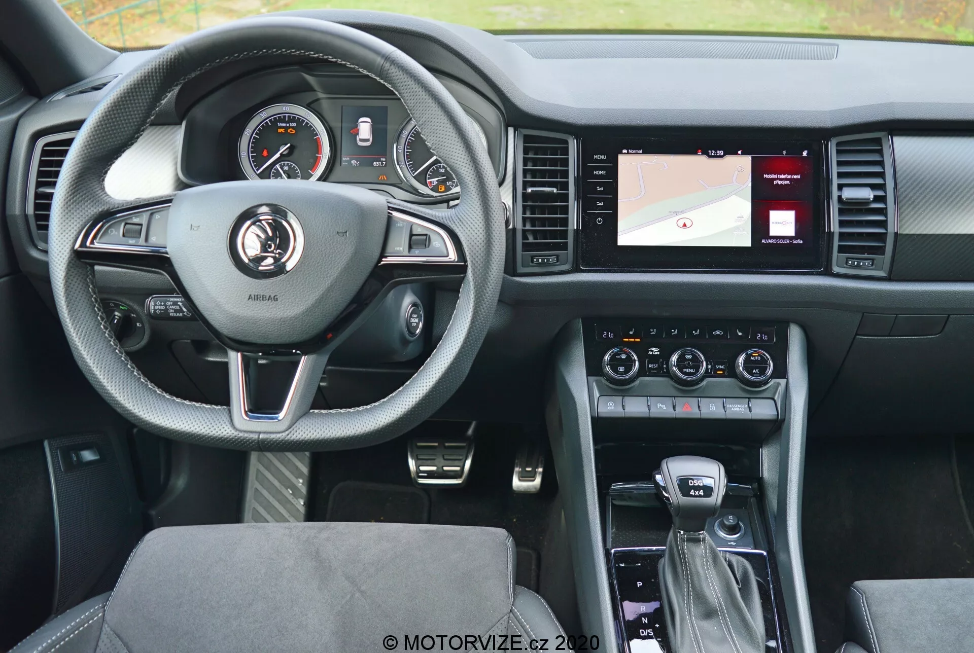 Vista interior de un Skoda Kodiaq 2017 desde el asiento trasero, destacando el volante de tres radios forrado en cuero con acentos plateados y botones de control para el control de crucero, el audio y la comunicación manos libres. El cuadro de instrumentos detrás del volante tiene diales analógicos y una pantalla digital. La consola central a la derecha incluye una pantalla con un mapa de navegación, controles de climatización y configuración de audio. La palanca de cambios sugiere una transmisión automática, con controladores adicionales para el sistema de infoentretenimiento y los modos de conducción. El diseño moderno busca un equilibrio entre comodidad y tecnología, con asientos de tela con patrón y una disposición bien iluminada y fácil de usar.