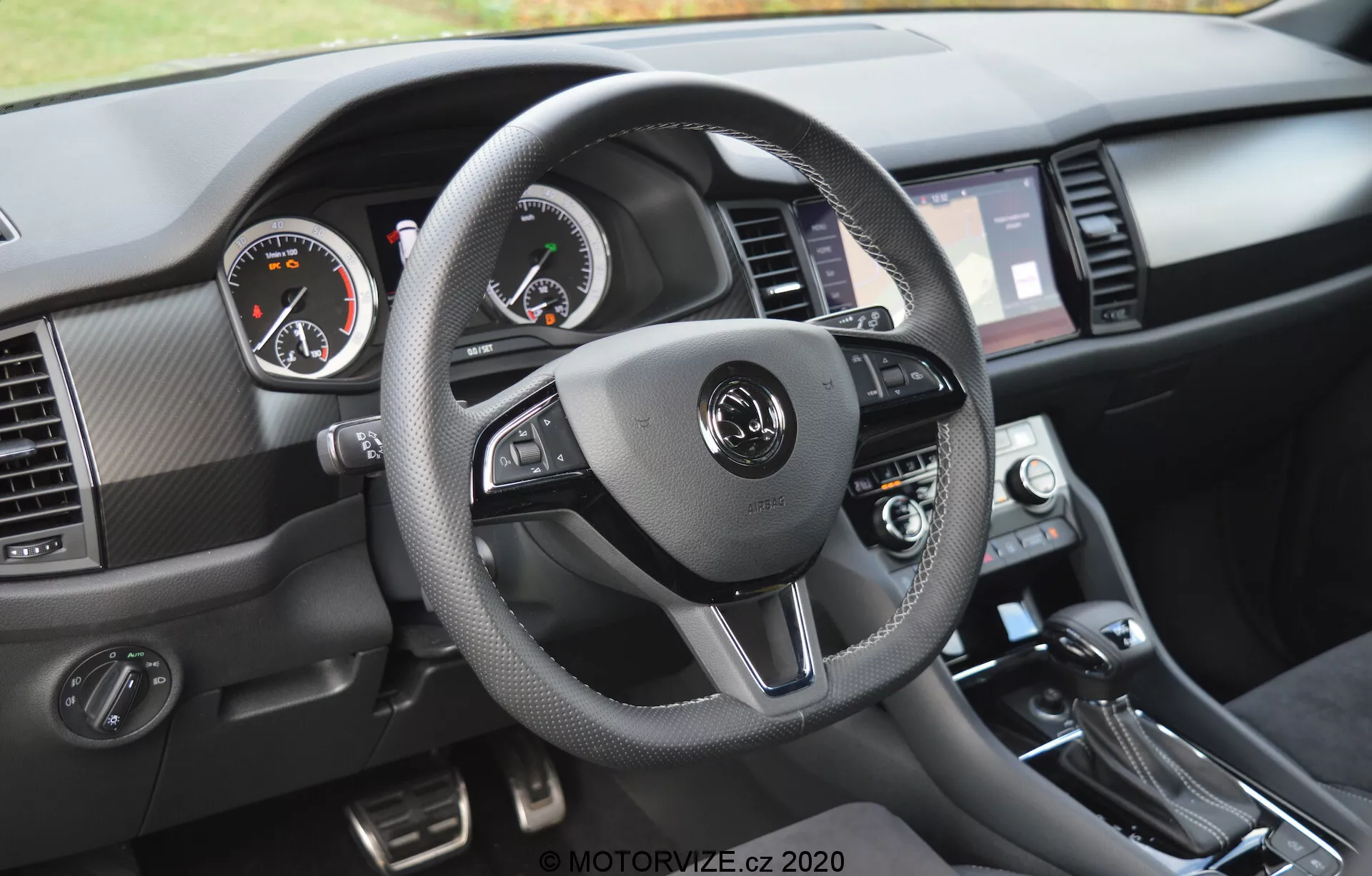 Vista interior de un Skoda Kodiaq 2017 centrada en la perspectiva del conductor del tablero. El volante de tres radios, envuelto en material que se asemeja al cuero con costuras contrastadas, cuenta con controles integrados para audio, comunicación manos libres y configuración de control de crucero. Los medidores analógicos, con el velocímetro a la izquierda y el tacómetro a la derecha, están complementados por una pantalla digital en el medio. La pantalla táctil de información y entretenimiento, flanqueada por rejillas de ventilación, diales de control de clima y palanca de cambios, son visibles, junto con materiales texturizados y acentos metálicos cepillados.