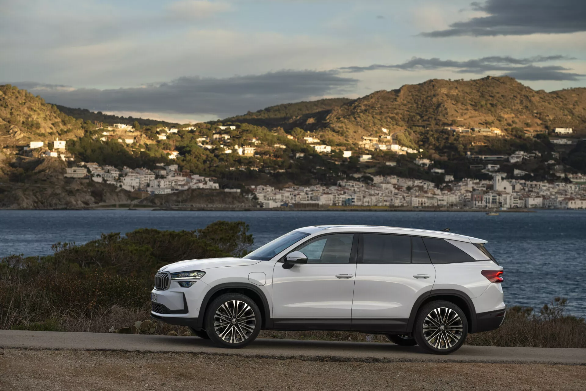 SUV moderno Skoda Kodiaq 2024 aparcado en una carretera costera con una vista escénica de una bahía y casas en la colina de fondo, con un diseño elegante, batalla larga, alta distancia al suelo, líneas de carrocería nítidas, manijas de puerta al ras, ventanas traseras tintadas, rieles de techo, grandes ruedas de aleación, faros LED, espejos laterales con señales de giro y techo solar panorámico, todo contra un acabado exterior blanco en contraste.