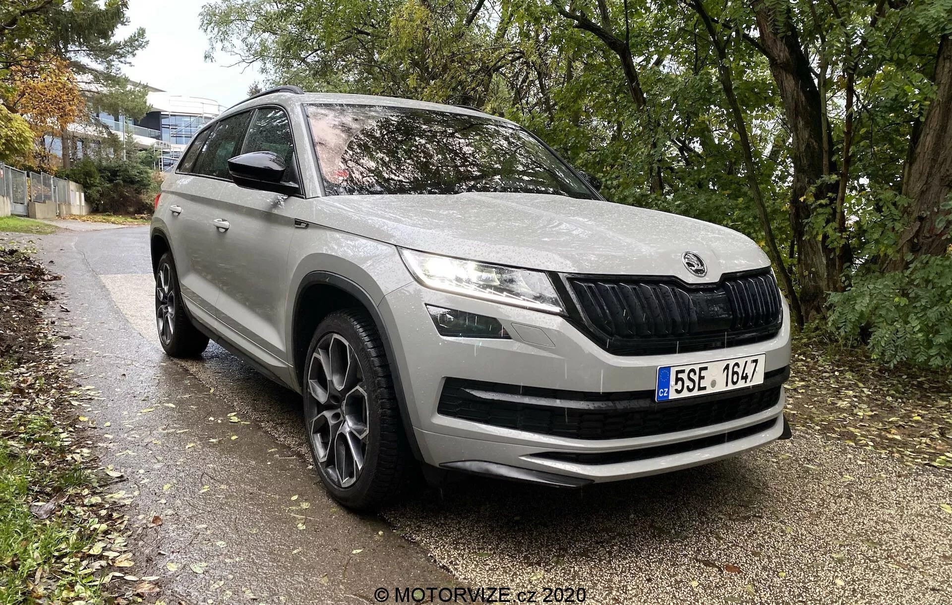 Skoda Kodiaq 2017 SUV moderno de una marca de autos europea, ángulo frontal de tres cuartos mostrando el perfil frontal y lateral hasta la mitad de la puerta trasera, estacionado junto a un camino bordeado de césped y árboles en un entorno urbano o suburbano. Las características incluyen una parrilla frontal característica con láminas verticales, faros LED elegantes, luces antiniebla en el parachoques, grandes entradas de aire en el parachoques delantero, líneas de contorno en el capó para aerodinámica, arcos de rueda prominentes con llantas de aleación de diseño multirradio, espejos laterales con intermitentes integrados, molduras protectoras en la parte inferior y pintura en general limpia y bien mantenida.