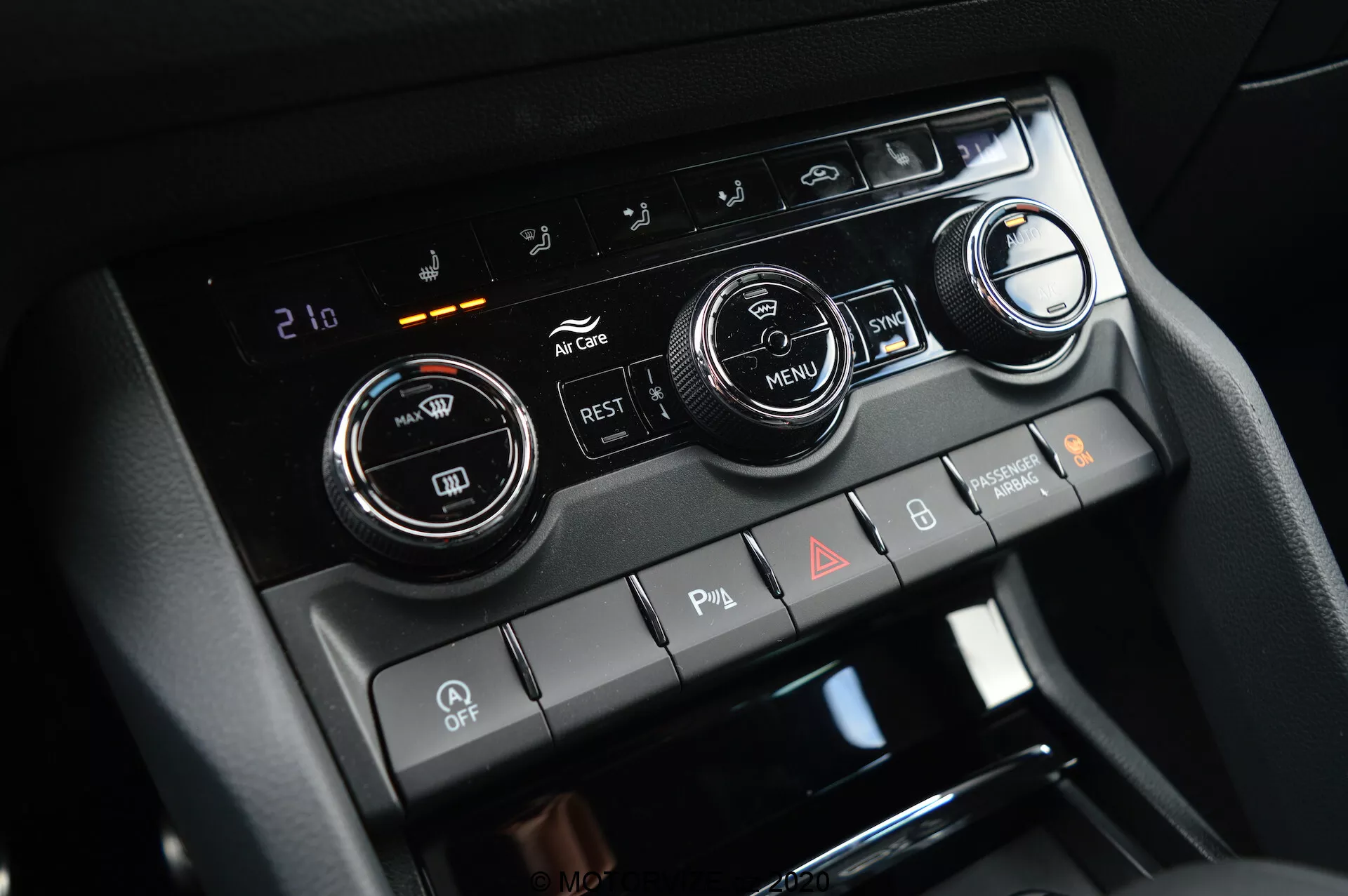 Vista en primer plano de la consola central del Škoda Kodiaq 2017, que destaca el sistema de control climático con dos grandes perillas de ajuste de temperatura y una pantalla digital ajustada a 21 grados Celsius. La imagen también muestra botones para aire acondicionado, recirculación, desempañadores de ventanas y un botón 'MENU'. Debajo hay interruptores para sensores de estacionamiento, desactivación de auto start/stop y luces de emergencia.