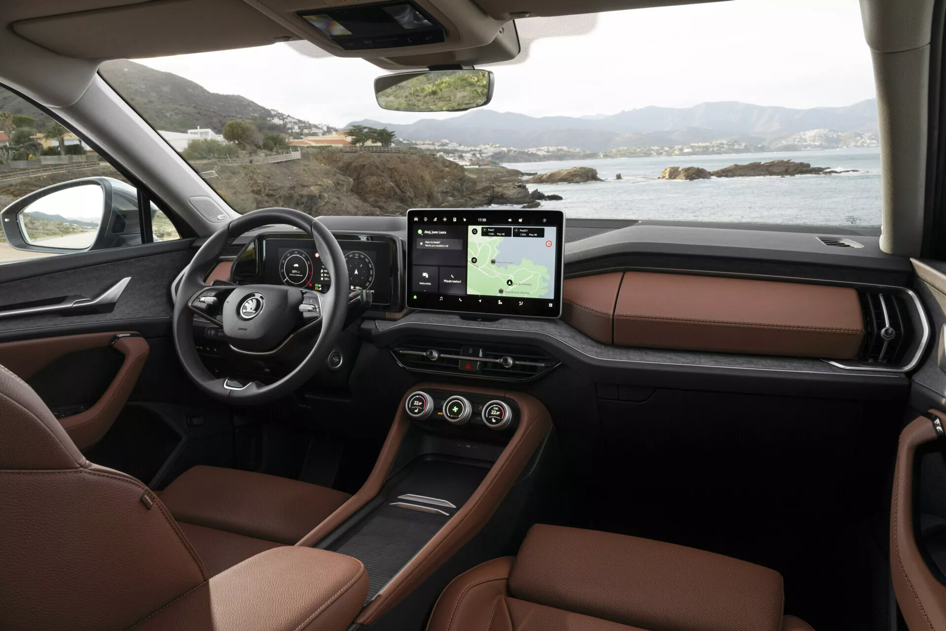 Vista interior de Skoda Kodiaq 2024 desde perspectiva del pasajero mostrando el tablero, volante multifunción con logo, cuadro de instrumentos digital, sistema de infotenimiento con pantalla táctil grande y GPS, asientos de cuero marrón lujosos y consola central elegante, ambientado contra un paisaje costero sereno afuera de las ventanas