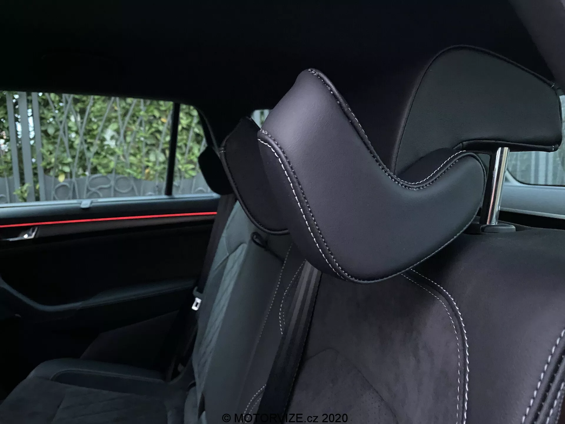 Vista interior de un Skoda Kodiaq 2017 centrada en un asiento de coche. La toma en ángulo enfatiza la parte del reposacabezas, tapizada en material negro, probablemente cuero o sintético de alta calidad, con un contorno distintivo para mayor comodidad. Las costuras precisas con hilo contrastante añaden detalles lujosos. En el fondo se revela una luz ambiental roja horizontal, destacando los elementos de diseño automotriz moderno. Las comodidades visibles para pasajeros traseros incluyen cinturones de seguridad y posiblemente un reposabrazos central, indicando la segunda fila de asientos.