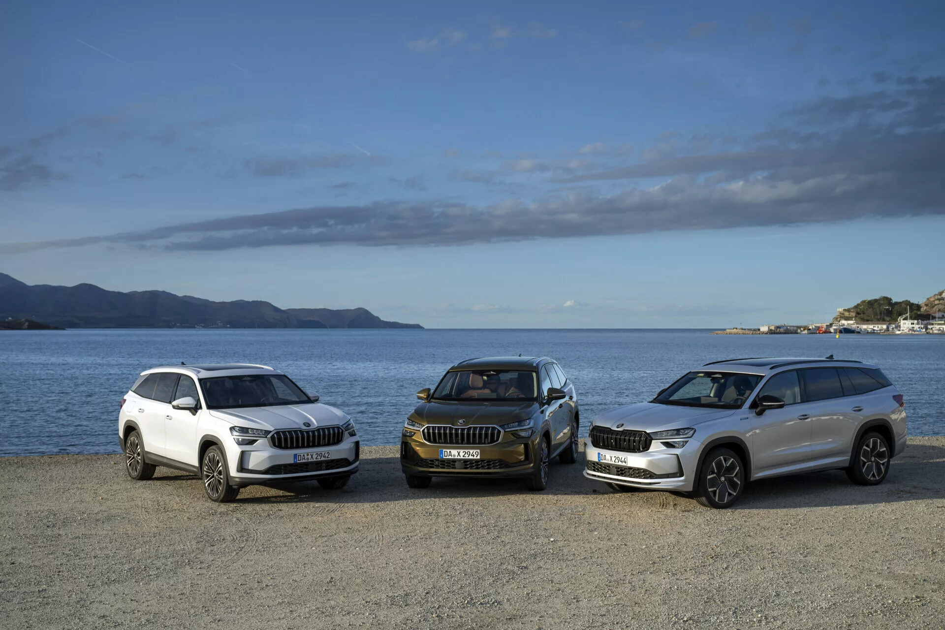 Gama de SUV Skoda Kodiaq 2024 en colores blanco y oscuro en vista frontal lateral, aparcados en un paseo marítimo con montañas y un cielo parcialmente nublado de fondo