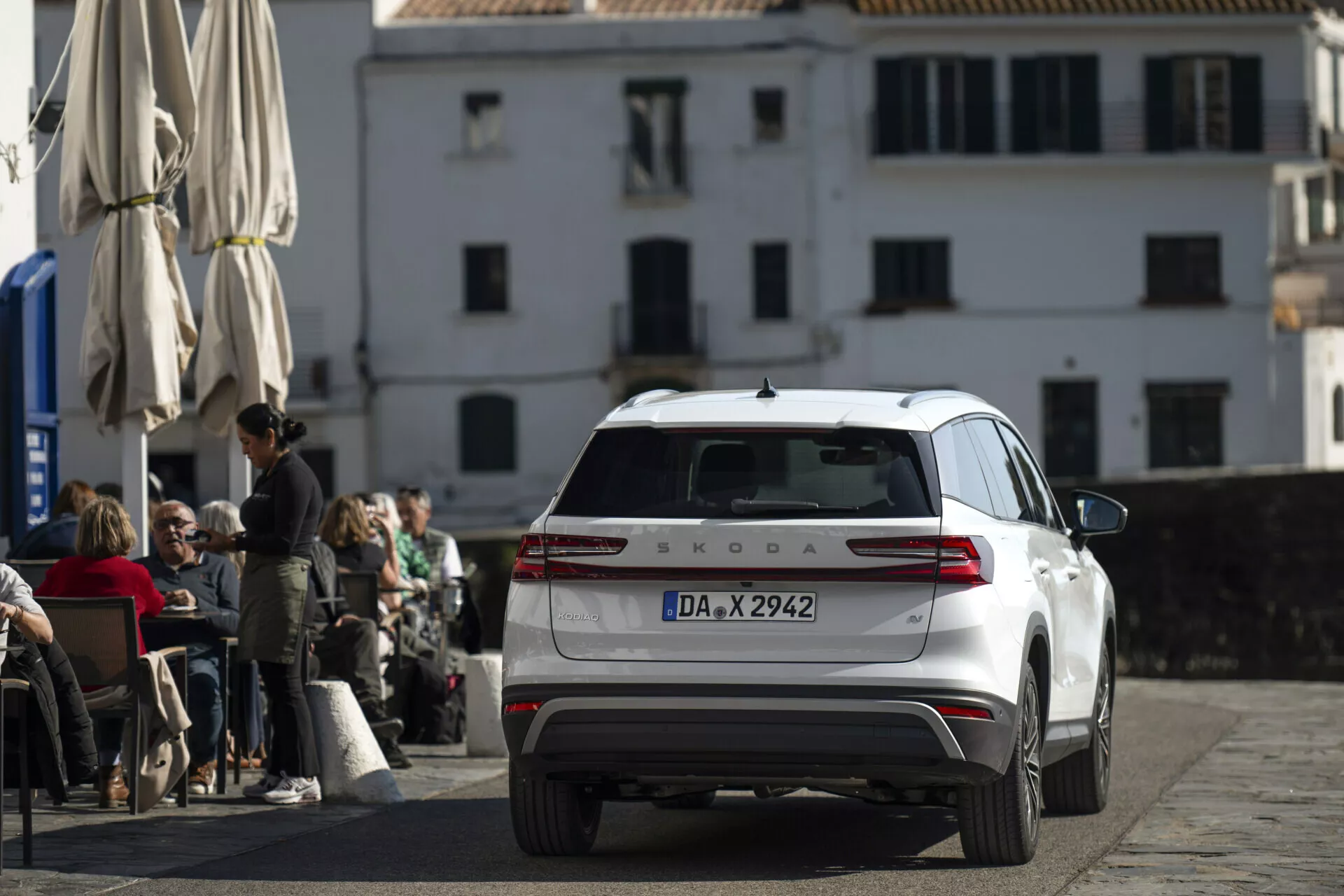 Škoda Kodiaq blanco del 2024 fotografiado desde un ángulo trasero y ligeramente elevado, mostrando la parte trasera completa, luces traseras, tapa del maletero, ventana trasera con limpiaparabrisas y techo con antena y barras, aparcado en una calle adoquinada frente a una cafetería con arquitectura europea clásica al fondo.