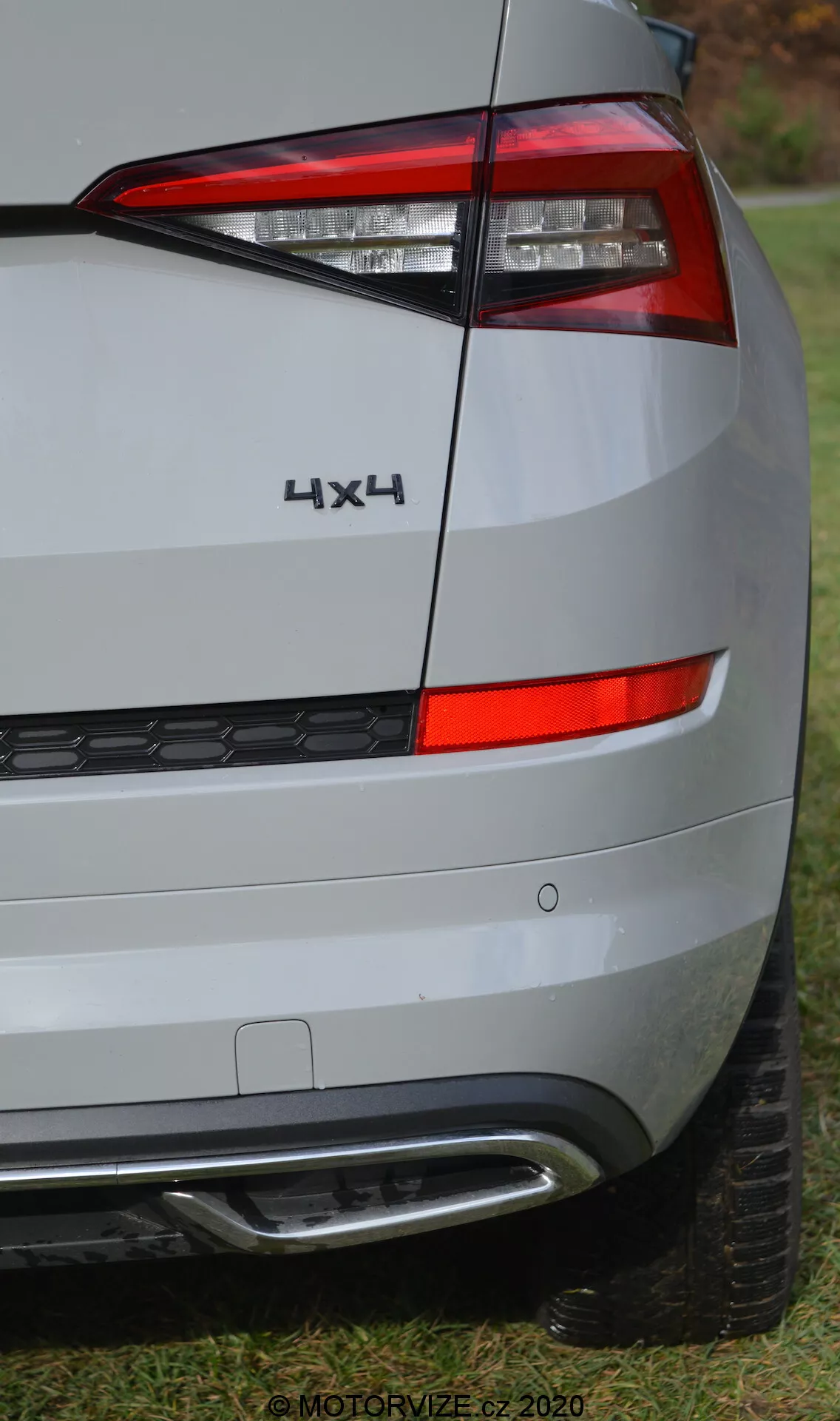 Vista parcial de cerca del rincón trasero del Skoda Kodiaq 2017, enfocándose en el diseño de las luces traseras, insignias y parachoques. El ángulo de la imagen enfatiza el lado derecho de la parte trasera del vehículo, mostrando un conjunto de luces traseras dispuestas horizontalmente con un diseño afilado y angular que rodea el lateral. Debajo de la luz trasera, se puede ver el panel trasero y el parachoques, de color plateado o gris claro, reflejando un ambiente apagado. El parachoques incluye una sección inferior de plástico negro con un patrón de panal y un elemento plateado o cromado, probablemente relacionado con asistencia de estacionamiento o estética. Una insignia '4X4' sobre la luz trasera indica tracción en las cuatro ruedas. La imagen de alta resolución revela texturas detalladas de la superficie, con una parte de la banda de rodadura visible al fondo, posicionada sobre césped. La marca de agua '© MOTORVIZE.cz 2020' indica que la imagen es para una publicación automotriz checa.