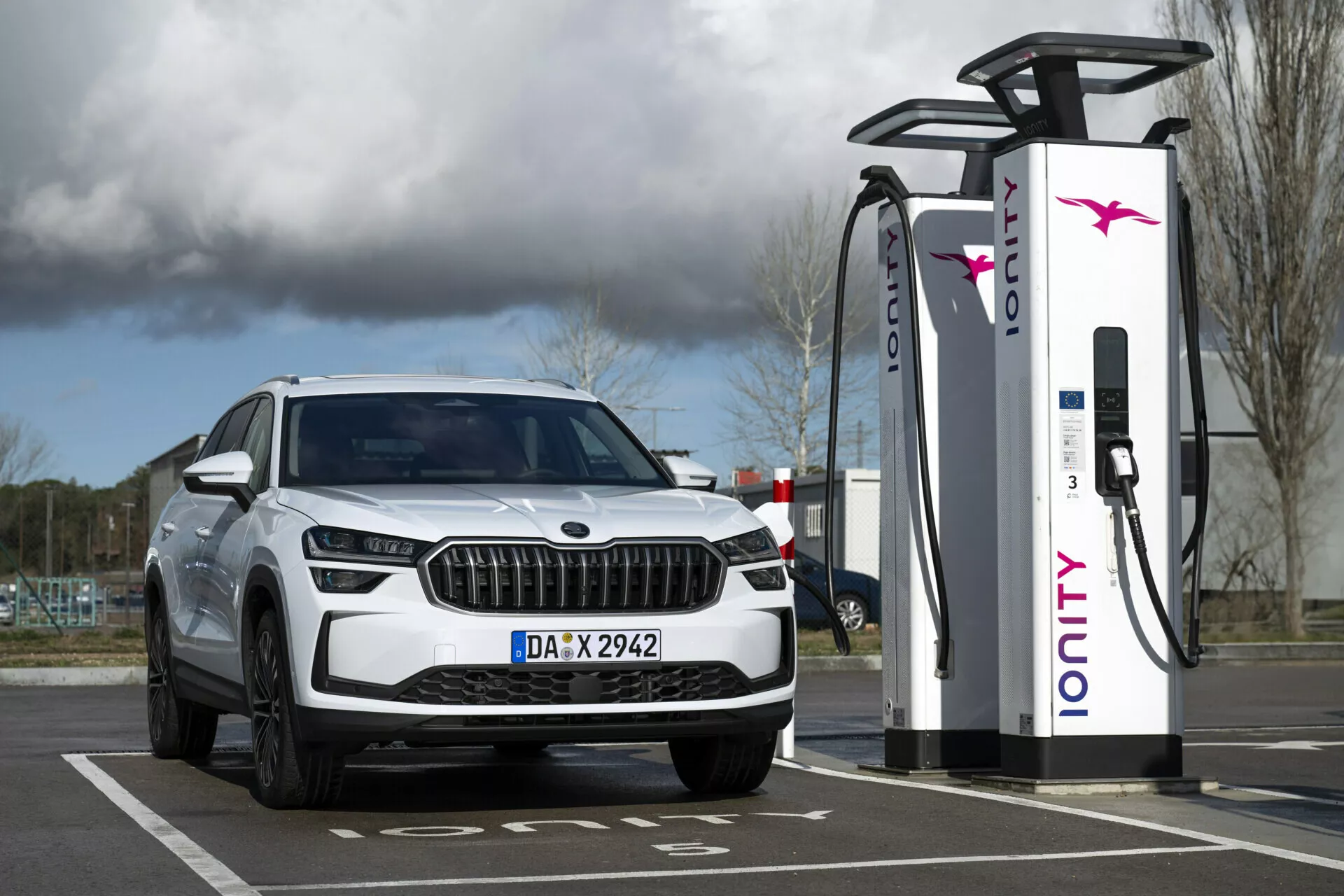 SUV blanco Skoda Kodiaq 2024 estacionado en una estación de carga eléctrica, mostrando la vista del cuarto delantero con faros LED y luces diurnas, enfatizando sus capacidades eléctricas o híbridas y diseño de parrilla lujoso.