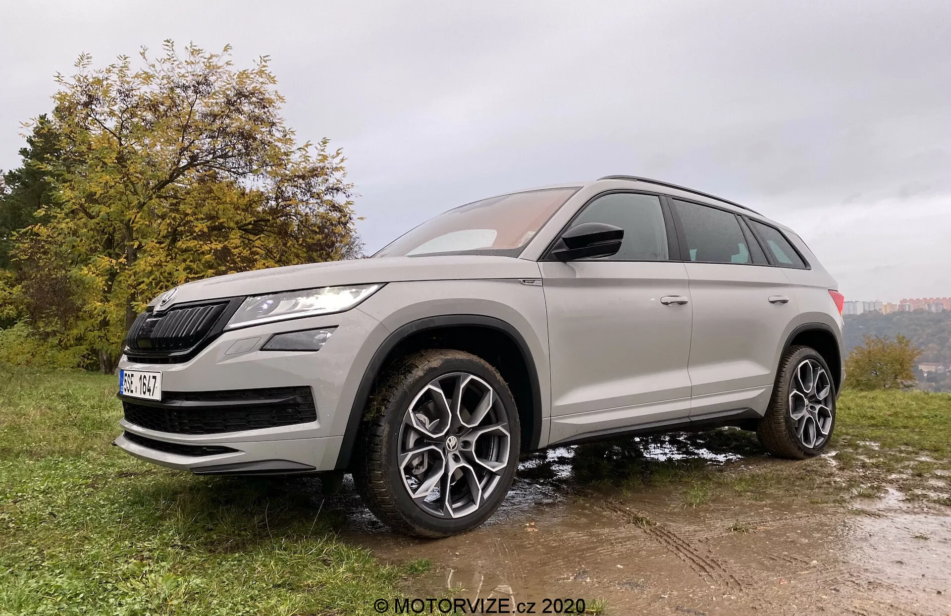 Vista delantera desde tres cuartos del SUV Škoda Kodiaq 2017 tomada desde un ángulo ligeramente bajo, enfatizando su altura y postura. El vehículo está estacionado en una superficie irregular de césped y barro, destacando sus capacidades todoterreno. El Škoda Kodiaq cuenta con una parrilla prominente y faros LED afilados. El diseño meticuloso incluye grandes ruedas de aleación de varios radios, gran altura del suelo y neumáticos para todas las estaciones. La carrocería está pintada en un color metálico neutro con ventanas tintadas, y los acentos cromados mejoran su atractivo premium. Las funcionalidades modernas se notan en los espejos laterales del color de la carrocería con indicadores de giro integrados y posiblemente sistemas de advertencia de ángulo muerto.