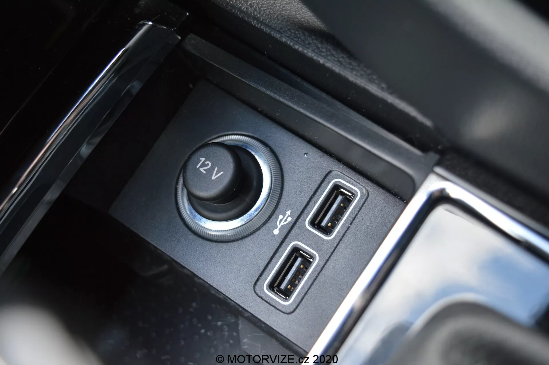 Primer plano del interior del Škoda Kodiaq 2017 enfocado en las funciones de suministro de energía y conectividad, incluyendo una toma de corriente de 12V y puertos USB duales, con un compartimento negro mate y acentos reflectantes brillantes.
