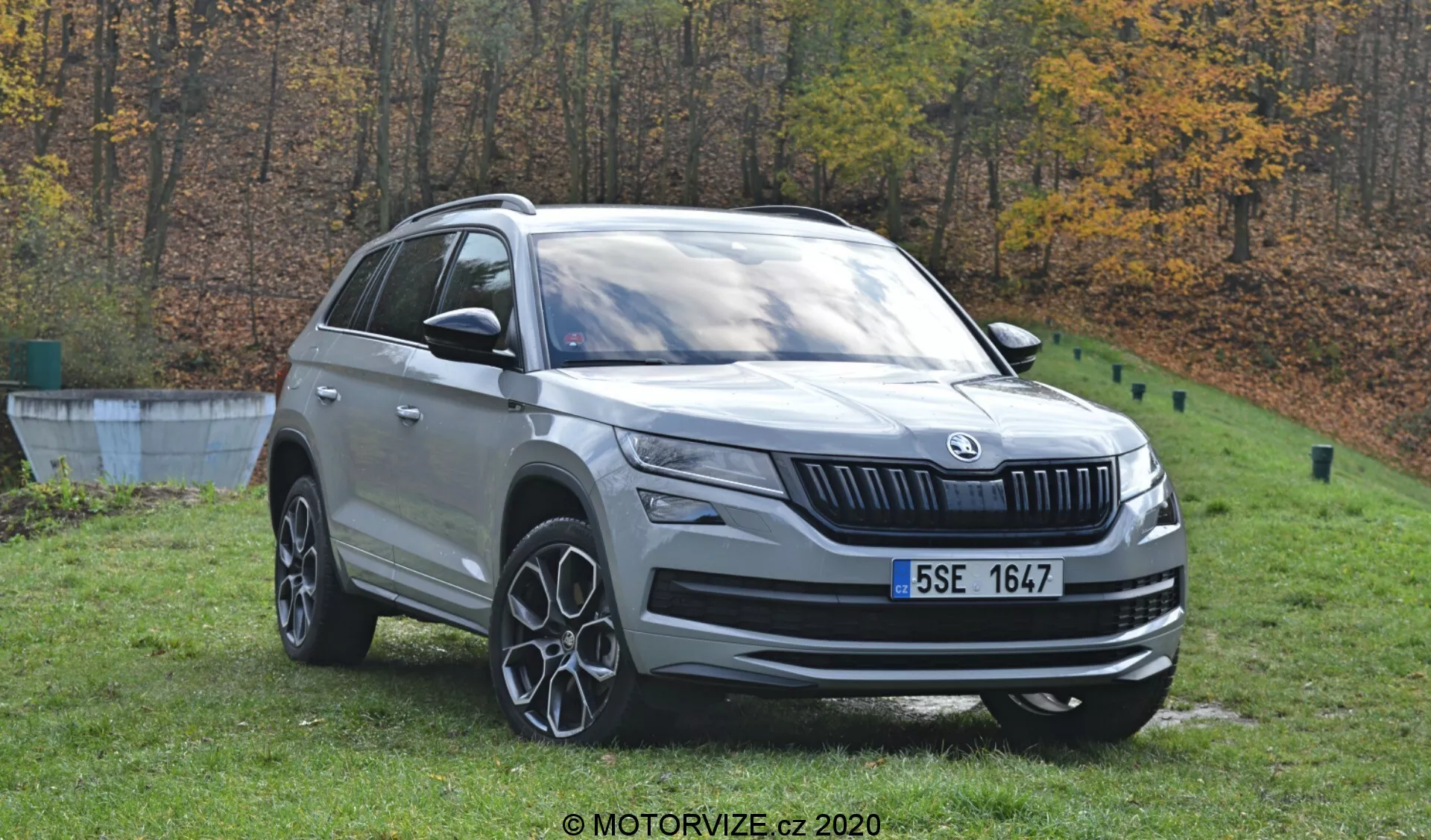 Una vista detallada del Škoda Kodiaq 2017, un SUV de color plateado, ubicado al aire libre con un fondo otoñal de árboles. La imagen diurna captura el vehículo desde un ángulo de tres cuartos delanteros, mostrando su perfil frontal y lateral. Se observan elementos de diseño distintivos, incluido el emblema de la marca en la parrilla frontal, faros LED, luces antiniebla, ruedas de aleación de varios radios, rieles de techo y espejos laterales con señales de giro integradas. El automóvil está posicionado sobre césped cerca de un borde de concreto, lo que indica un entorno similar a un parque.