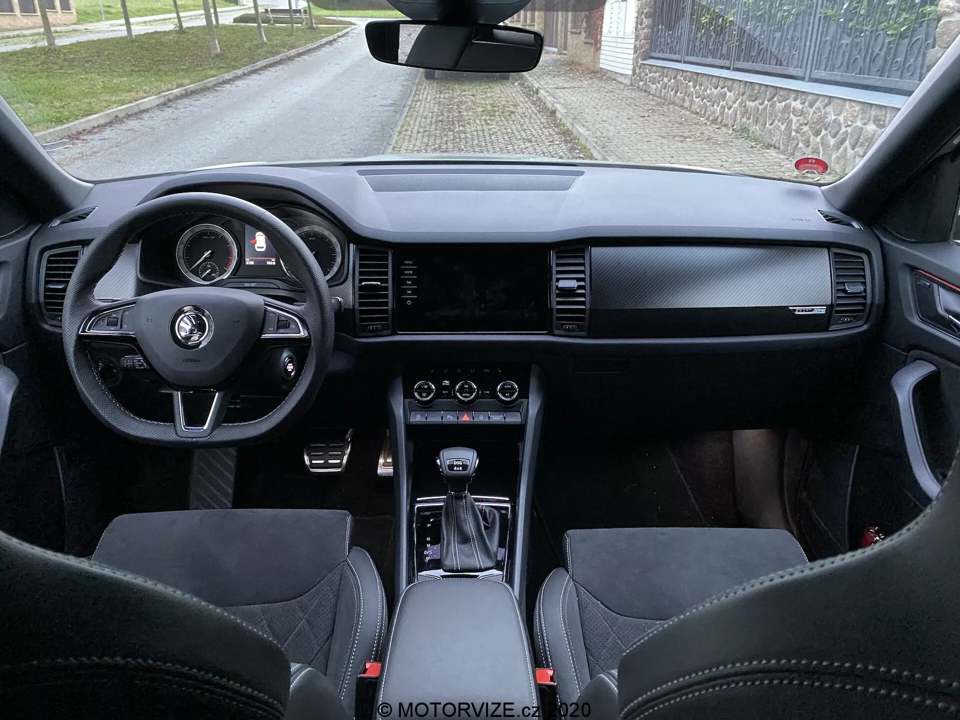 Vista interior del Skoda Kodiaq 2017 desde el espacio del asiento trasero, mostrando el tablero de instrumentos, volante con botones multifunción, cuadro de instrumentos digital, pantalla de infoentretenimiento ancha, controles del HVAC, selector de cambios de transmisión automática, materiales de cuero premium y asientos deportivos con reposacabezas integrados.
