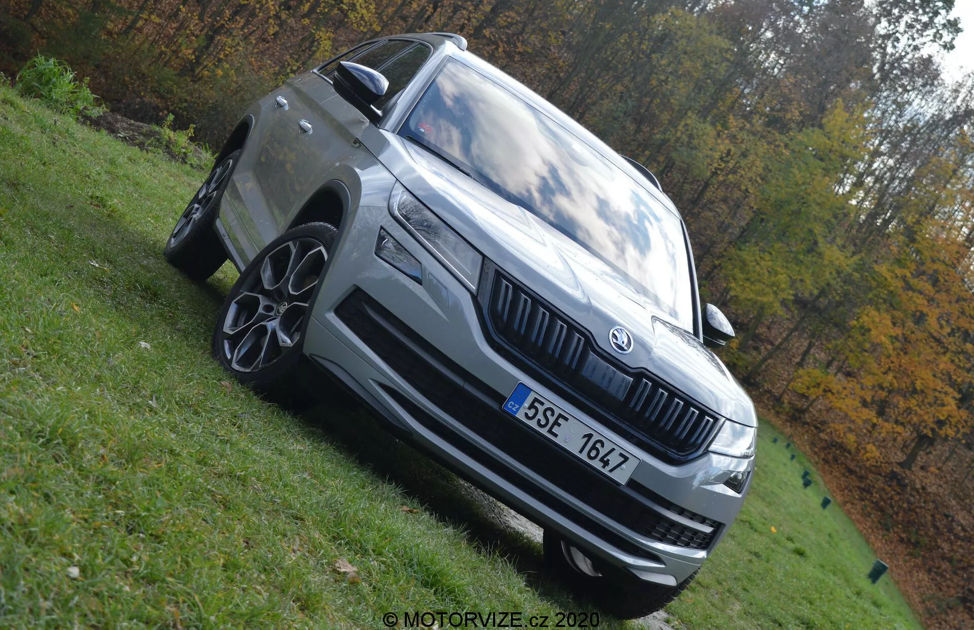 Fotografía en ángulo bajo de un Skoda Kodiaq 2017 destacando su estatura dominante. El coche se muestra desde el cuarto delantero izquierdo con características notables como faros LED, parrilla prominente con el emblema de Skoda, llantas de aleación multipalo, parachoques delantero con entradas de aire, espejo lateral con señal de giro integrada y líneas de diseño exterior distintivas. El vehículo es de color gris claro o plateado y está aparcado en un área de césped bajo luz natural del día. El fondo otoñal borroso acentúa la presencia del vehículo.
