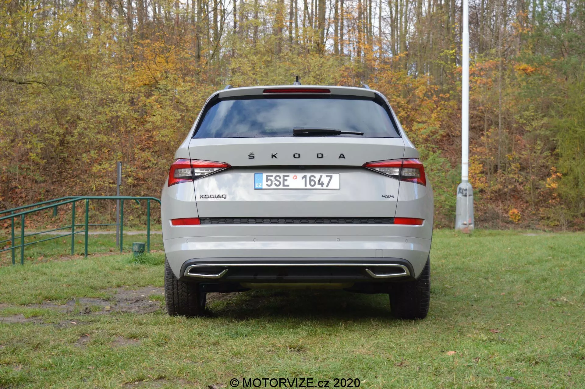 Imagen trasera de un vehículo Škoda Kodiaq 2017 en un entorno al aire libre. La marca 'ŠKODA' y el nombre del modelo 'KODIAQ' se muestran de manera prominente en la tapa del maletero. Matrícula de estilo europeo, luces traseras modernas con luz antiniebla trasera y luces de reversa integradas, parachoques trasero visible con sensores de estacionamiento y parabrisas trasero con limpiaparabrisas son claramente visibles. Los elementos reflectantes en la parte inferior del parachoques mejoran la visibilidad. El coche parece estar aparcado en una zona de césped ligeramente embarrada, lo que sugiere capacidad todoterreno.