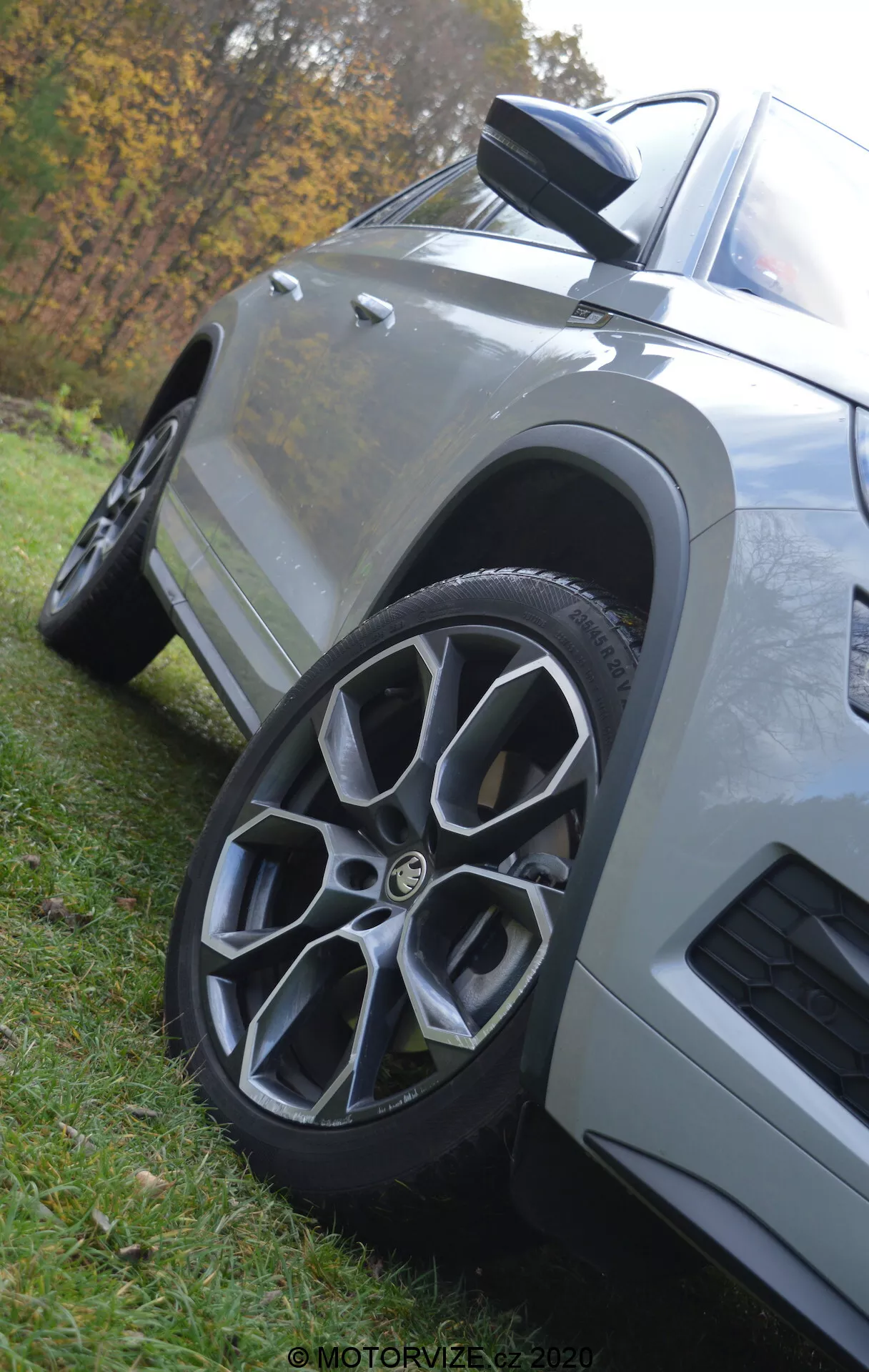 Vista de cerca y a baja altura del Škoda Kodiaq 2017 destacando la rueda delantera y la aleta. El coche, aparcado en el césped, tiene una llanta de aleación multirradio, espejo lateral aerodinámico con indicador integrado, paneles de carrocería angulares con acabado de pintura metalizada y una ventilación en el guardabarros delantero, todo con un fondo natural desenfocado.