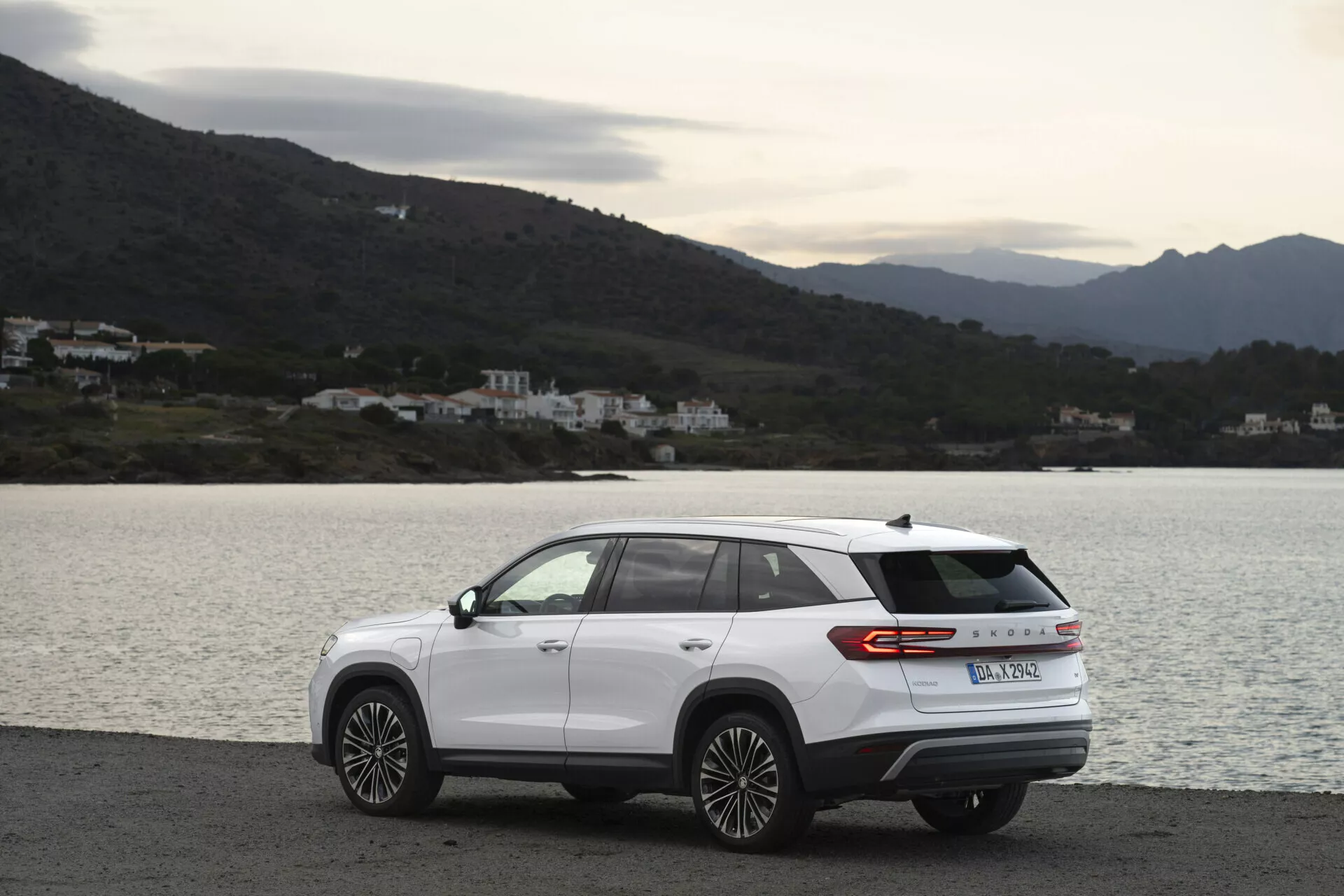 SUV moderno Skoda Kodiaq 2024 aparcado lateralmente en una carretera costera escénica, con diseño aerodinámico, tira de luz trasera y ruedas de aleación de múltiples radios, en un día nublado con iluminación natural difusa, con un fondo de agua, colinas y edificios