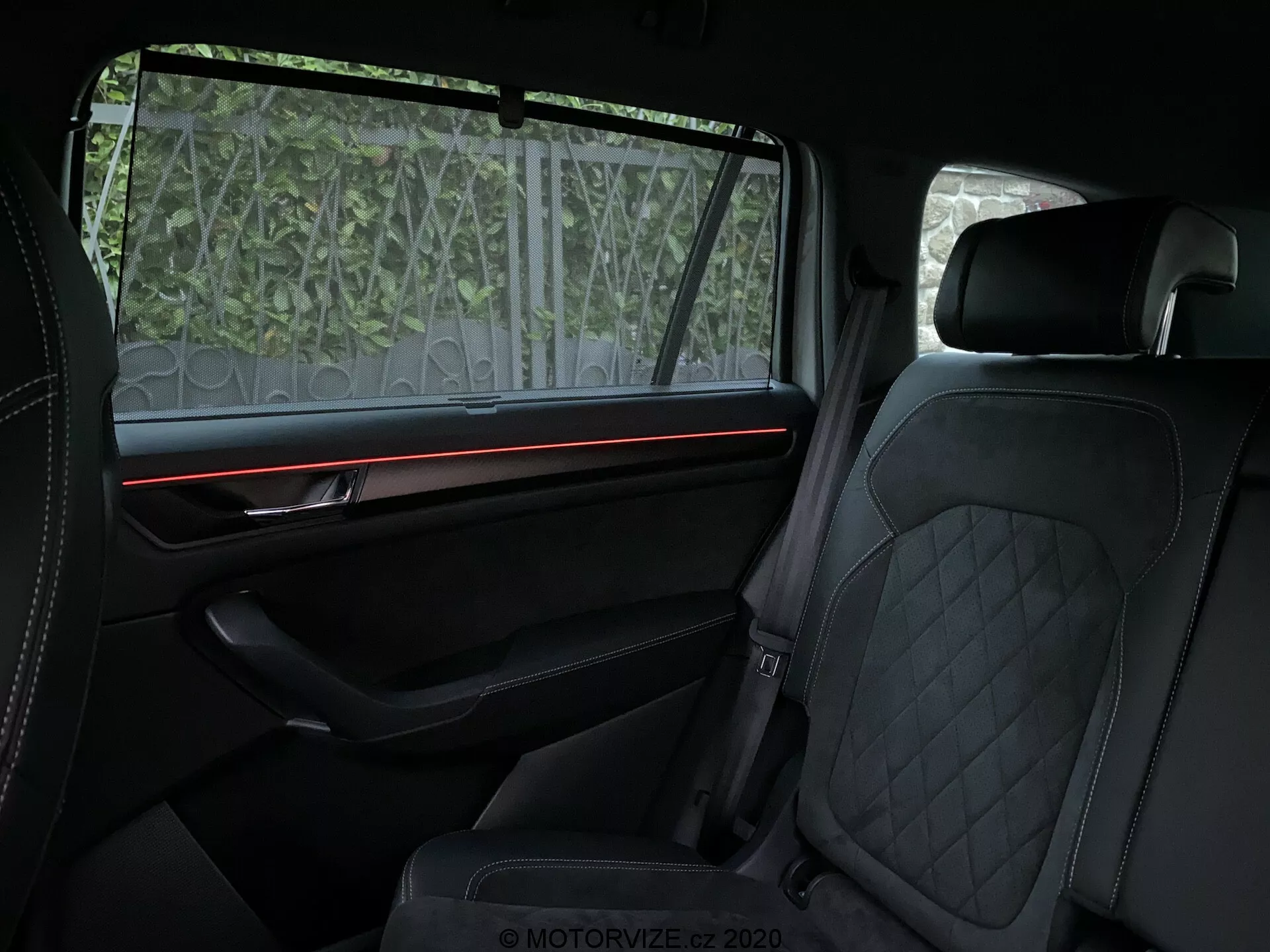 Vista interior del Skoda Kodiaq 2017 enfocada en el compartimiento trasero de pasajeros, mostrando características de lujo como tapicería de cuero con costura en diamante en los asientos delanteros y traseros, tira de iluminación ambiental roja en el panel de la puerta bien equipado con adorno premium, y una pantalla de protección solar parcialmente translúcida en la ventana. El énfasis está en la comodidad y elementos de diseño de alta gama.