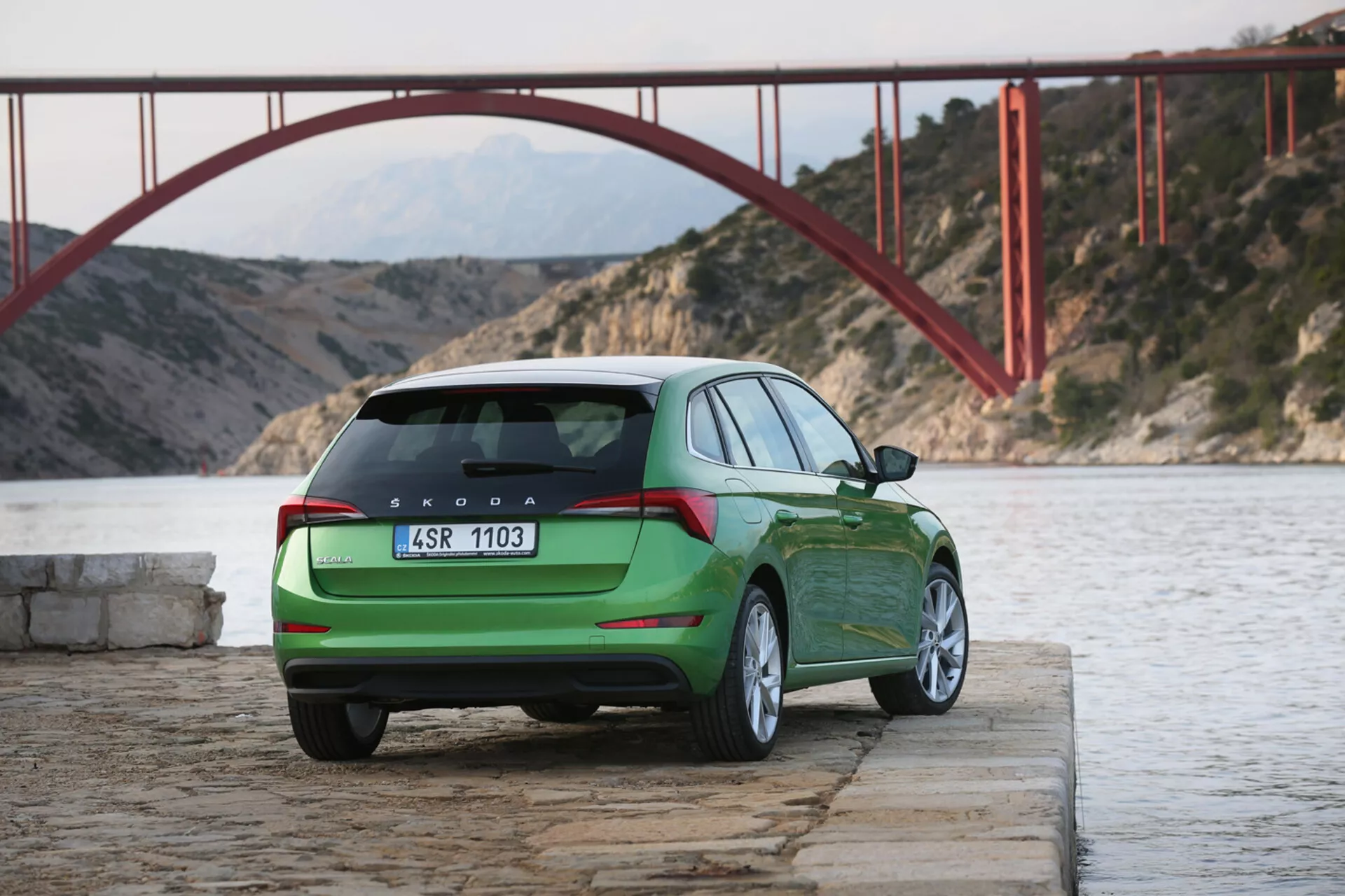 Skoda Scala verde del 2019 estacionado junto al agua con vista del perfil trasero y lateral, mostrando las luces traseras, portón trasero, logotipo y designación del modelo con un puente rojo y montañas al fondo