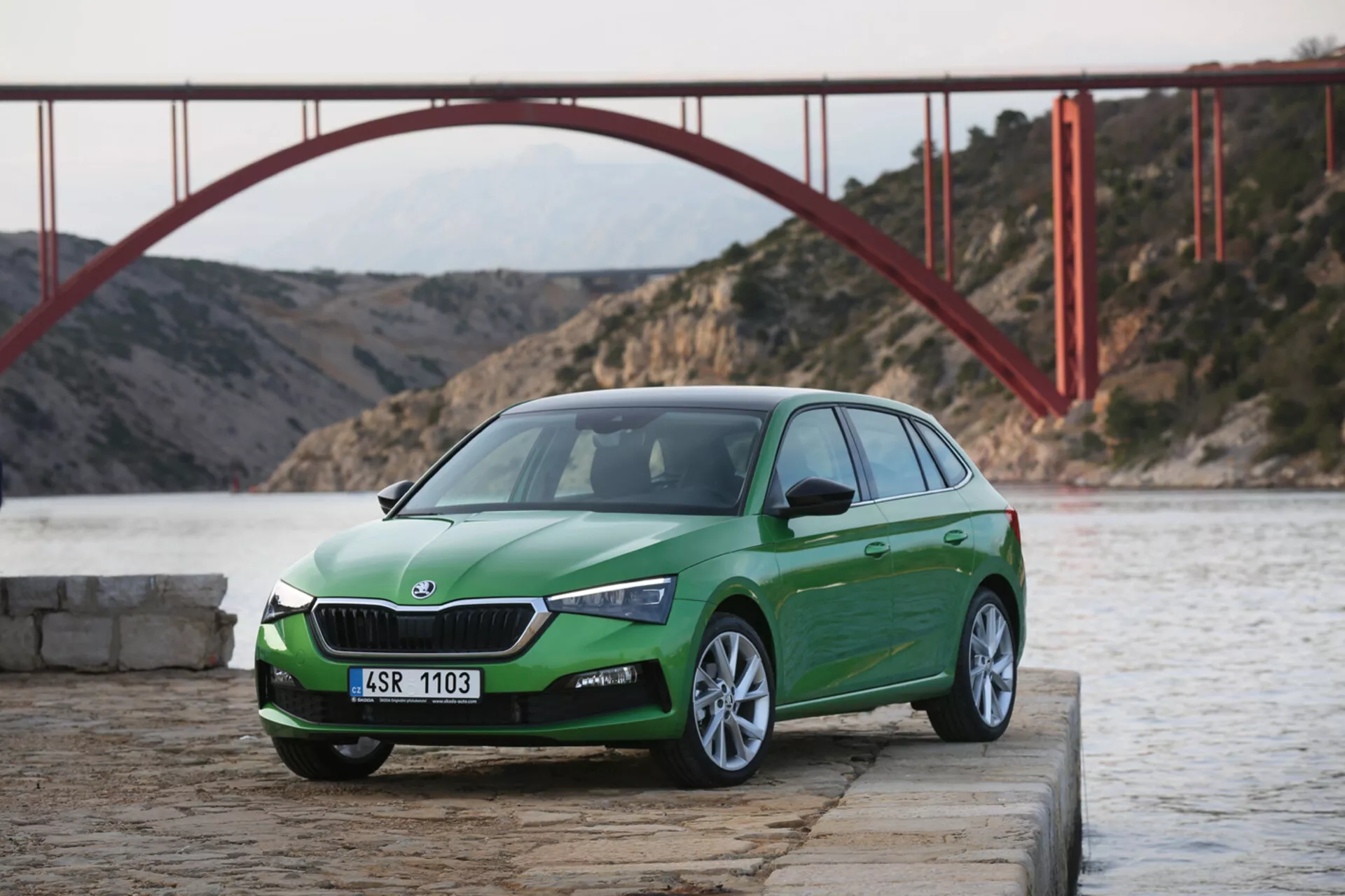 Vista angular frontal tres cuartos de un hatchback Skoda Scala 2019 en verde, sobre concreto cerca del agua con puente rojo de fondo durante el día, mostrando diseño y características