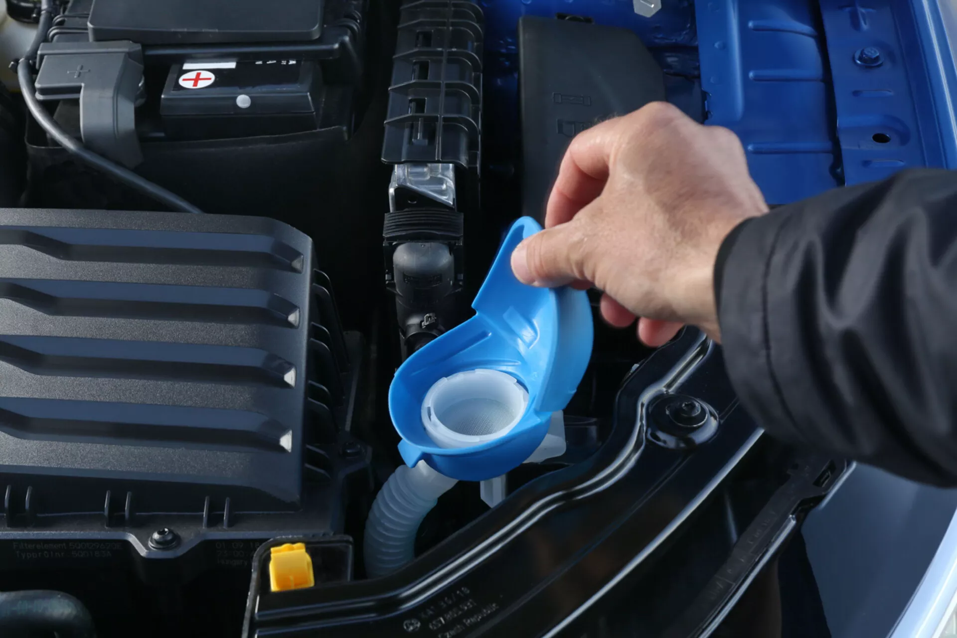 Vista detallada del compartimento del motor de Skoda Scala 2019 enfocando en el rellenado del líquido limpiaparabrisas con un embudo azul, mostrando la batería, la cubierta del motor y la estructura del vehículo