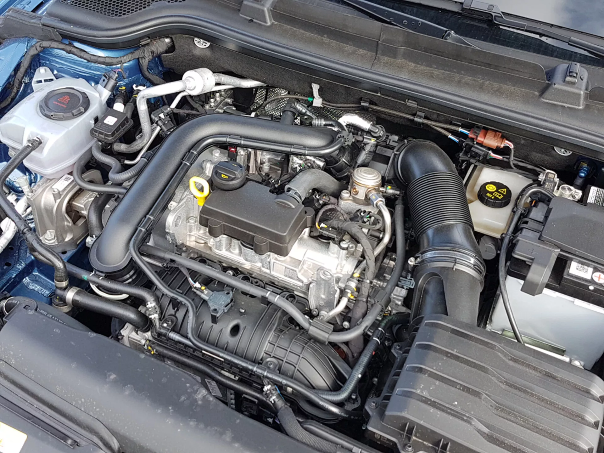 Vista detallada del compartimento del motor de un Skoda Scala 2019 con el capó abierto, mostrando el bloque del motor y varios componentes como el contenedor de refrigerante, la carcasa del filtro de aire y la batería, indicativo de ingeniería automotriz y atención al mantenimiento.