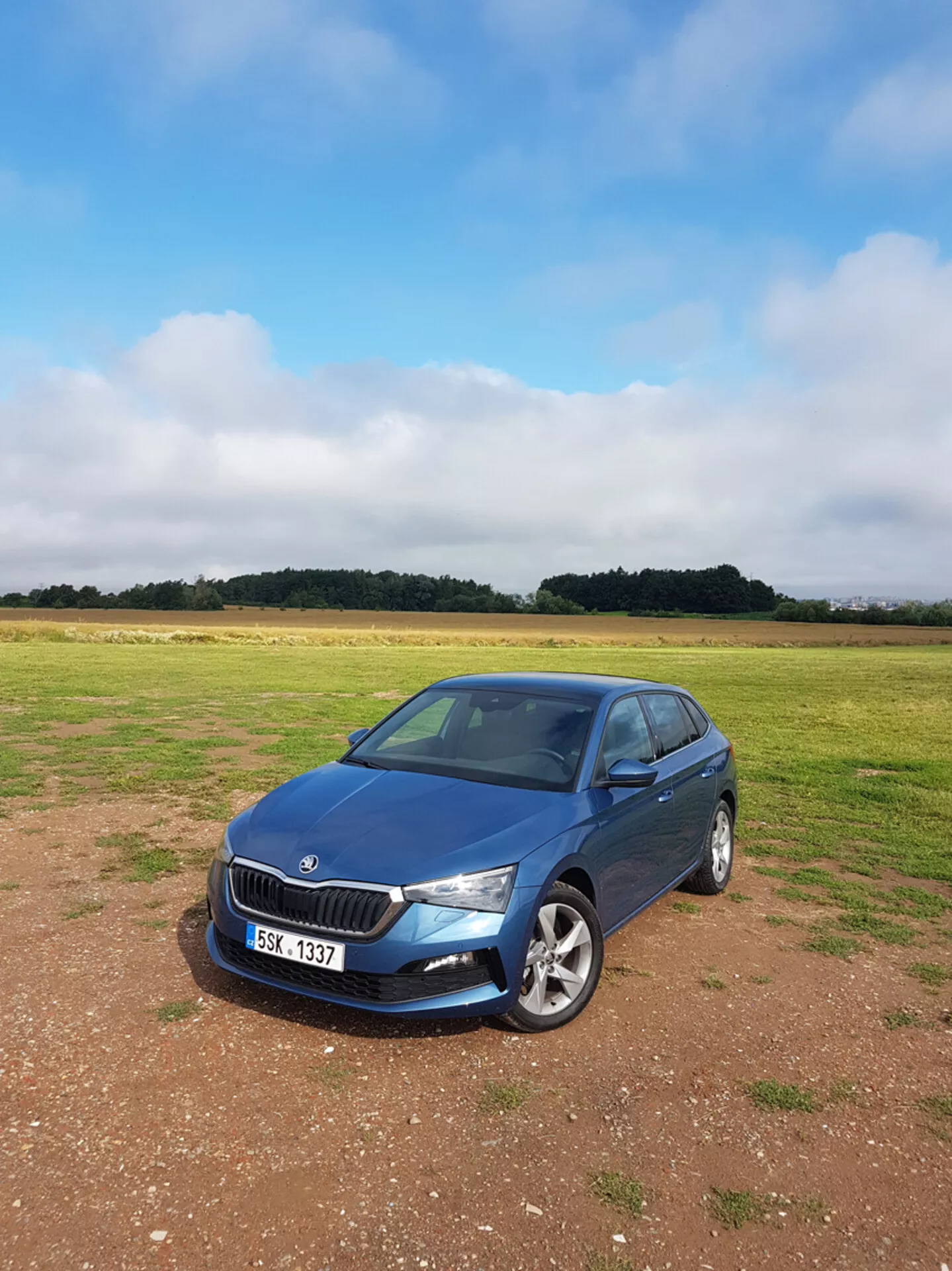 Sedán Skoda Scala 2019 en azul, aparcado sobre césped con cielo nublado, mostrando parrilla frontal, faros y perfil lateral del pasajero con fondo de campo