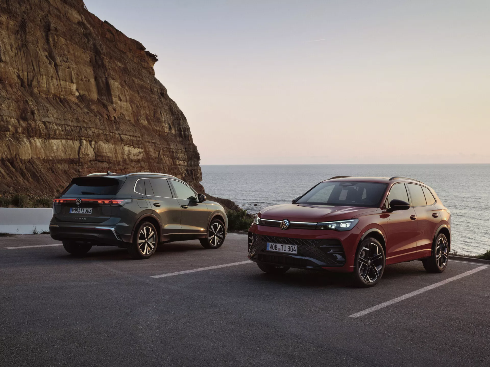 SUVs de lujo en un escenario costero pintoresco durante la puesta del sol, con un modelo gris con luces traseras LED y elementos de diseño deportivo y un modelo burdeos con una parrilla frontal orientada al rendimiento y sistemas avanzados de asistencia al conductor