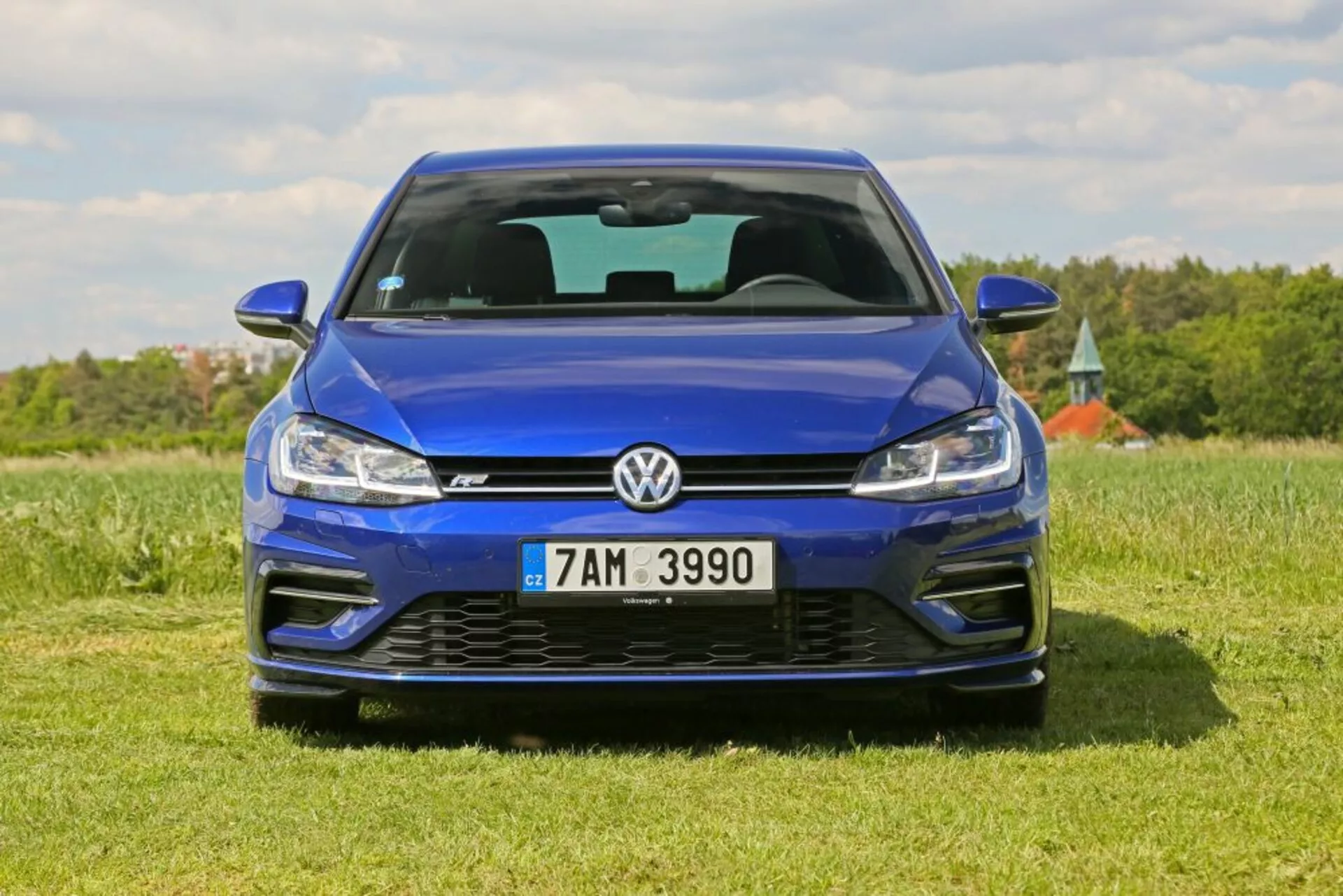 Vista frontal de un Volkswagen Golf azul aparcado en un campo de hierba bajo un cielo despejado, destacando su acabado metálico, diseño de rejilla, tecnología de faros LED y parachoques delantero deportivo incluidos los conductos de aire y el emblema.